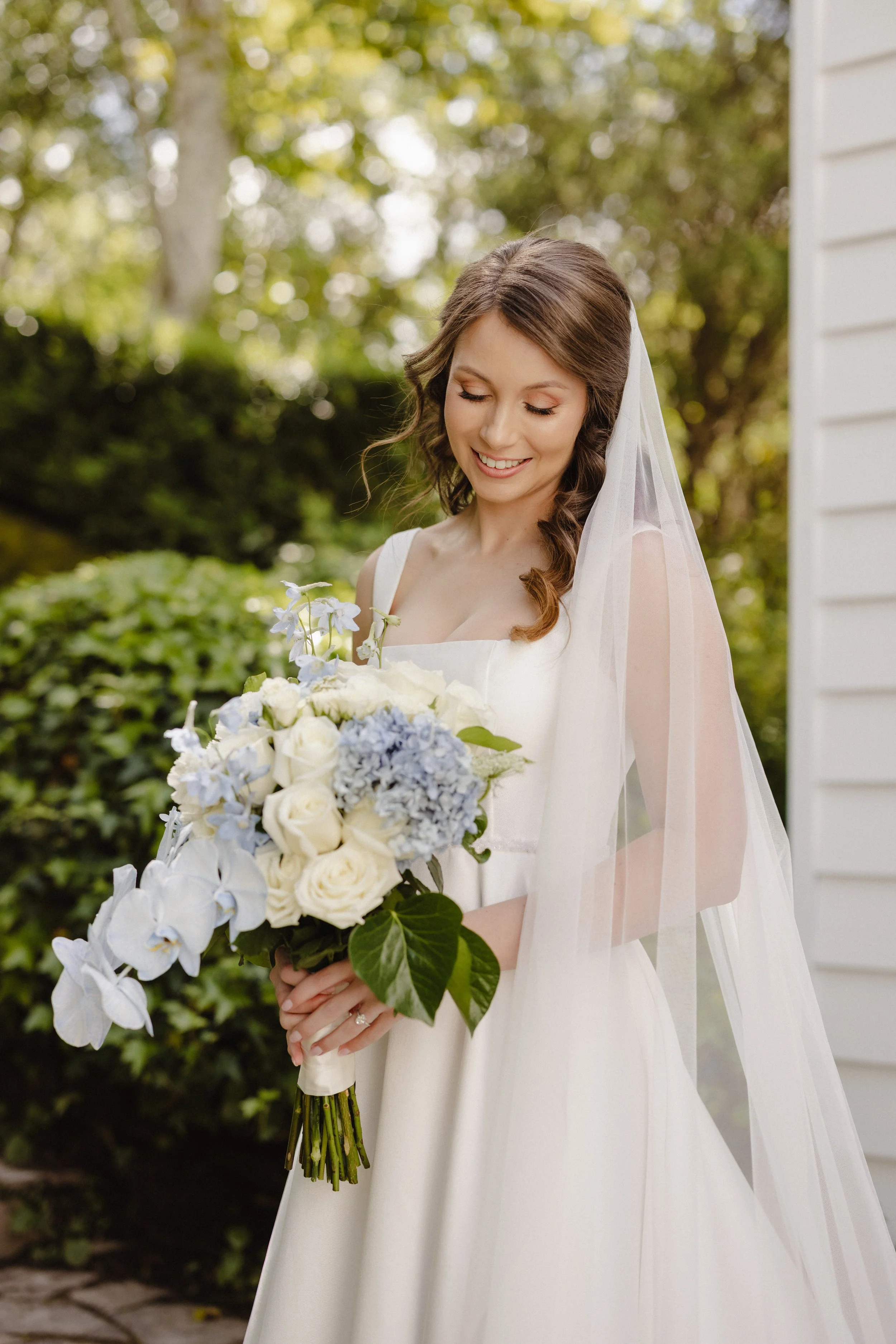 A bride in a white wedding dress holding a bouquet of white and blue flowers, standing outdoors with greenery in the background.