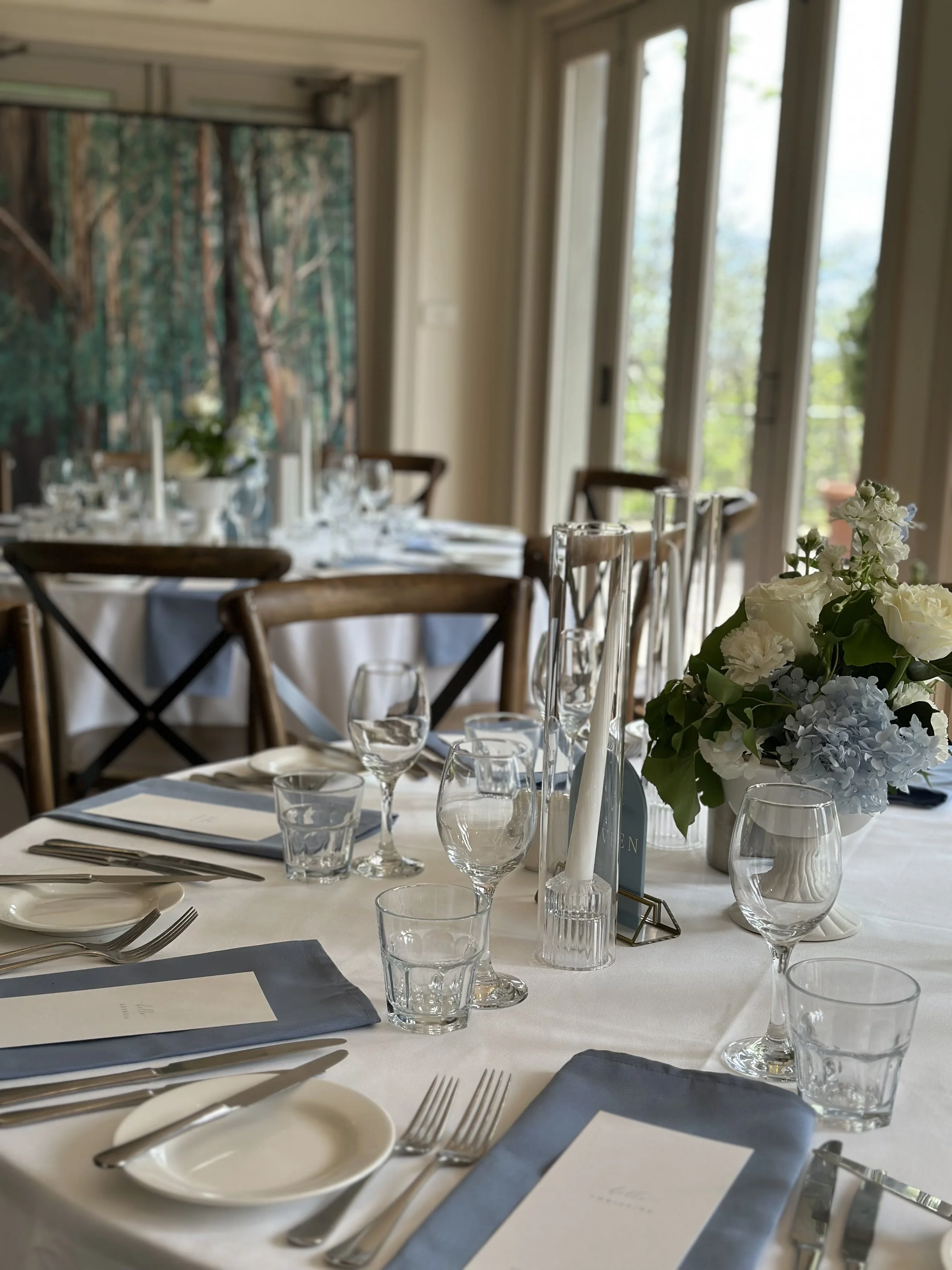 A formal dining table set with glasses, plates, silverware, napkins, and a floral centerpiece in a well-lit room with large windows and a forest-themed painting in the background.