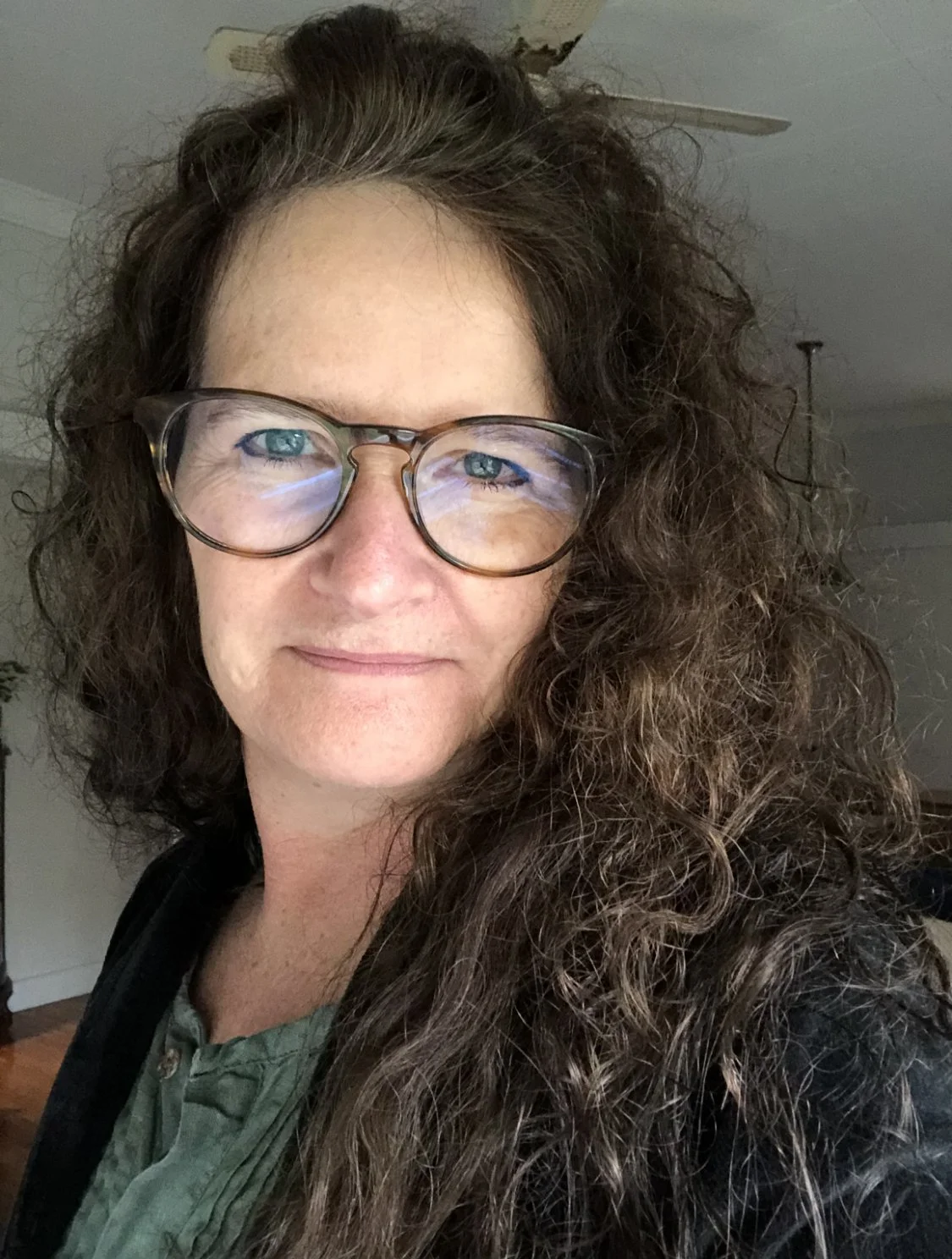 A woman with curly brown hair and glasses smiling at the camera indoors.