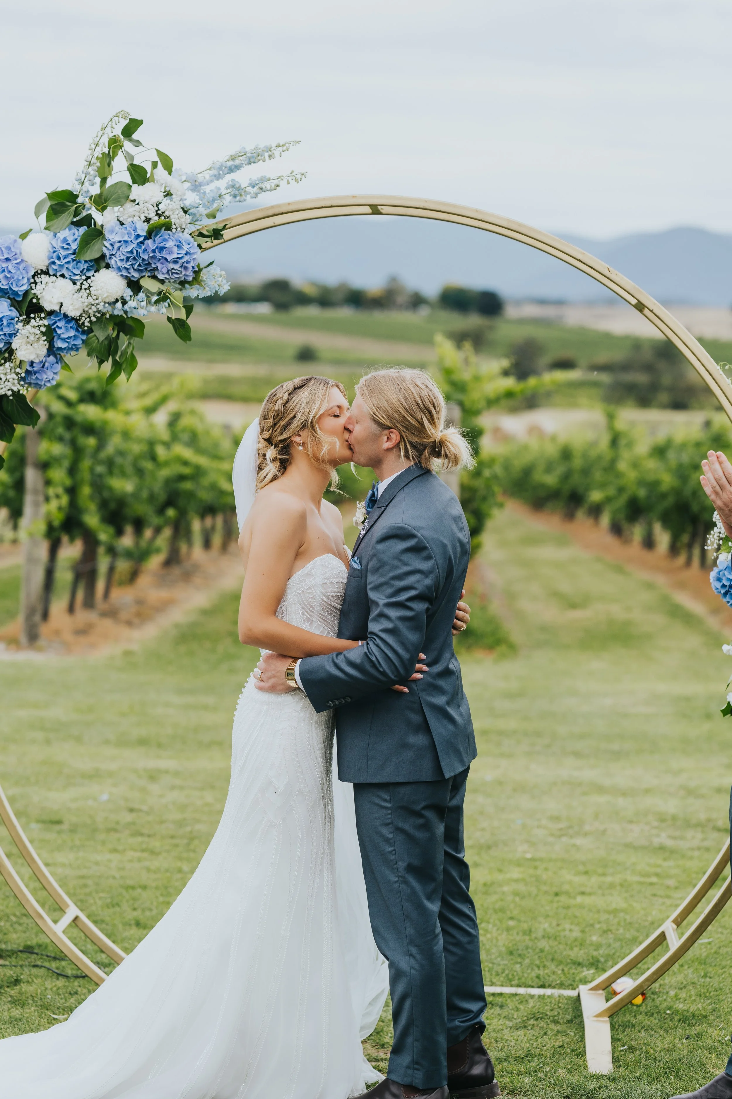 A bride and groom kiss at their outdoor wedding ceremony, standing under a circular floral arch with blue and white flowers, on a grassy field with vineyard rows and hills in the background.
