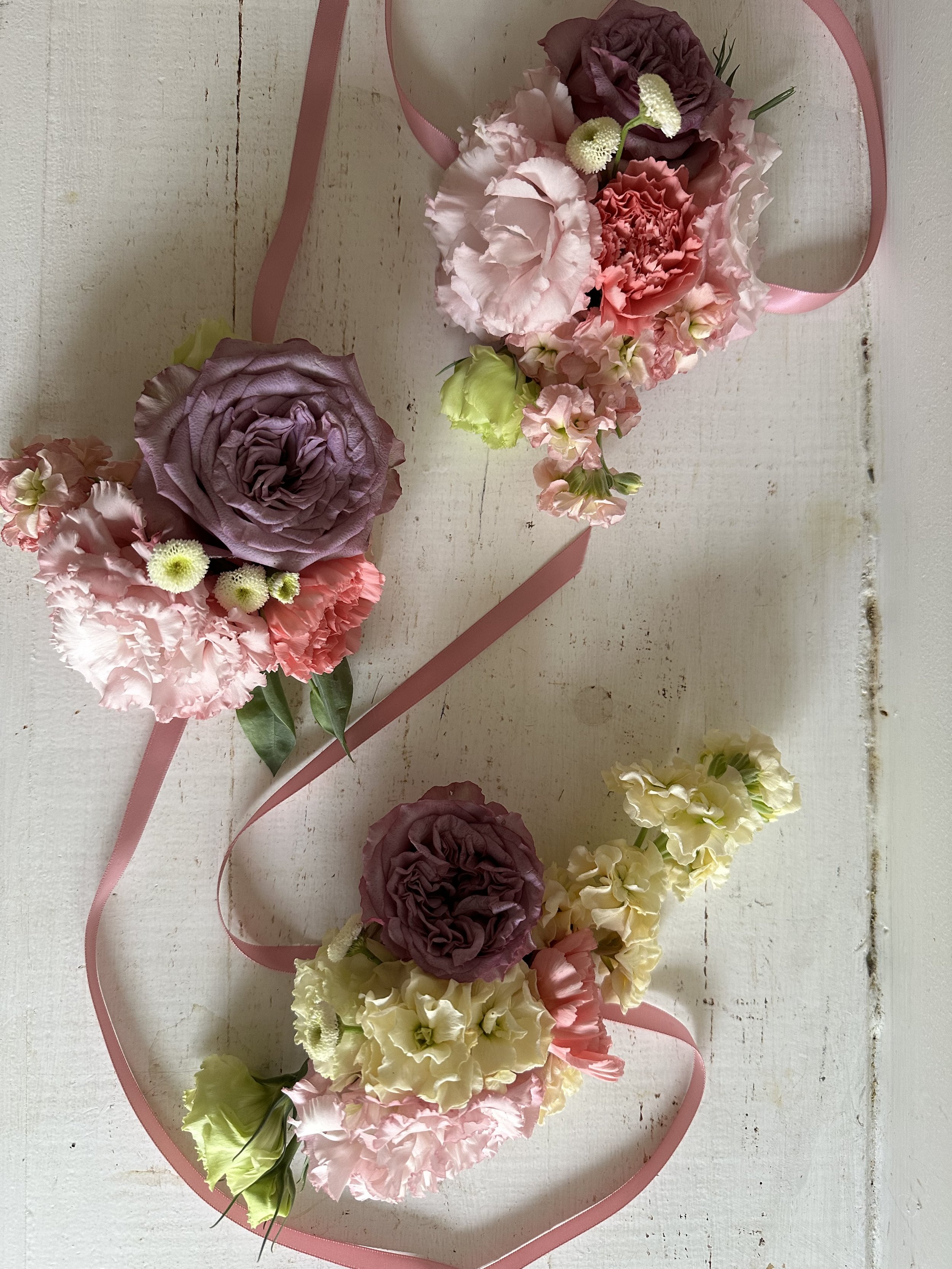 Colorful floral wreath with pink, purple, and white flowers on a white wooden background, accented with pink ribbon.