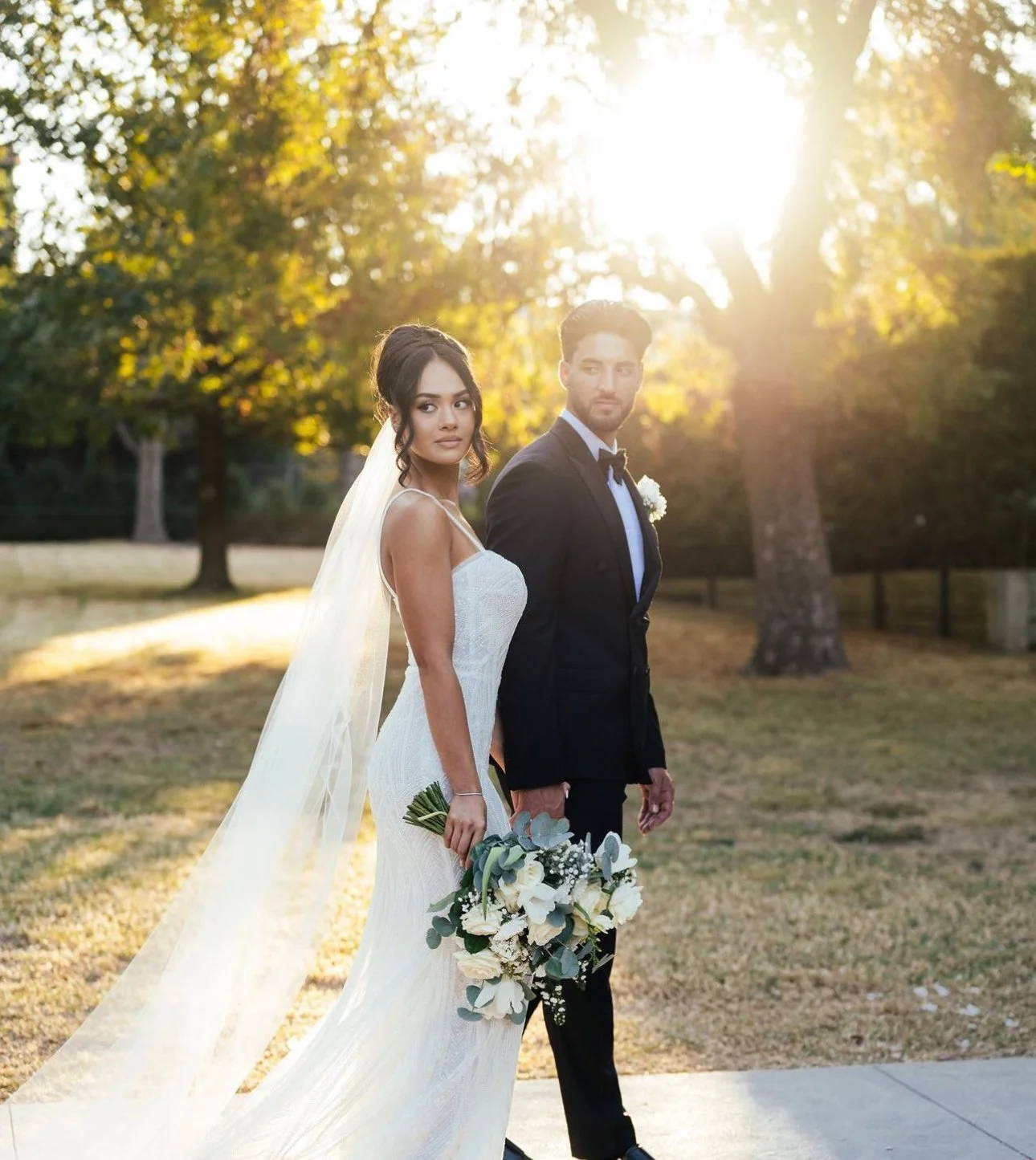 A bride and groom standing outdoors during sunset, dressed in wedding attire with trees in the background.
