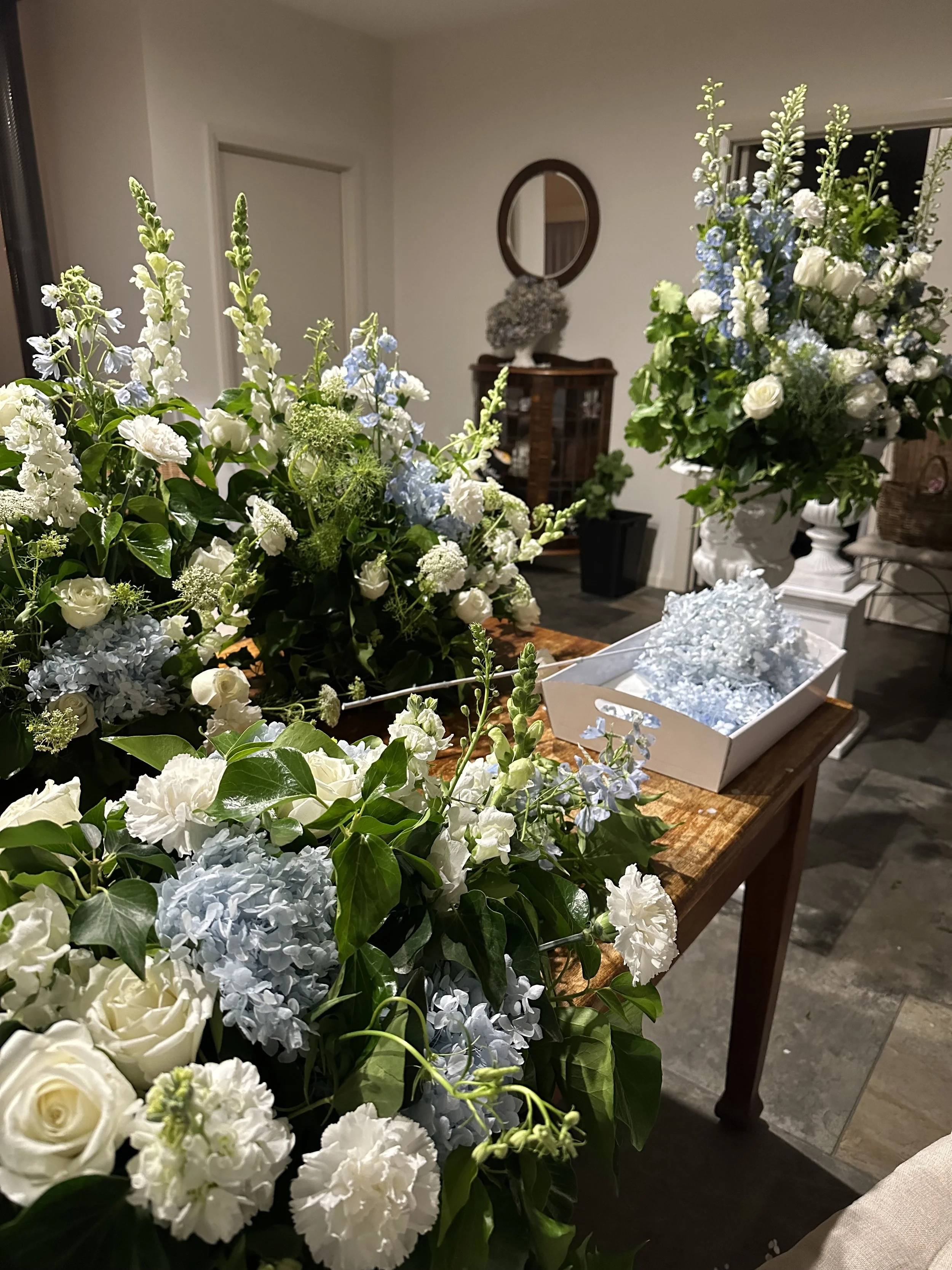 A room decorated with large white and blue floral arrangements on a wooden table, with a mirror and shelves in the background.