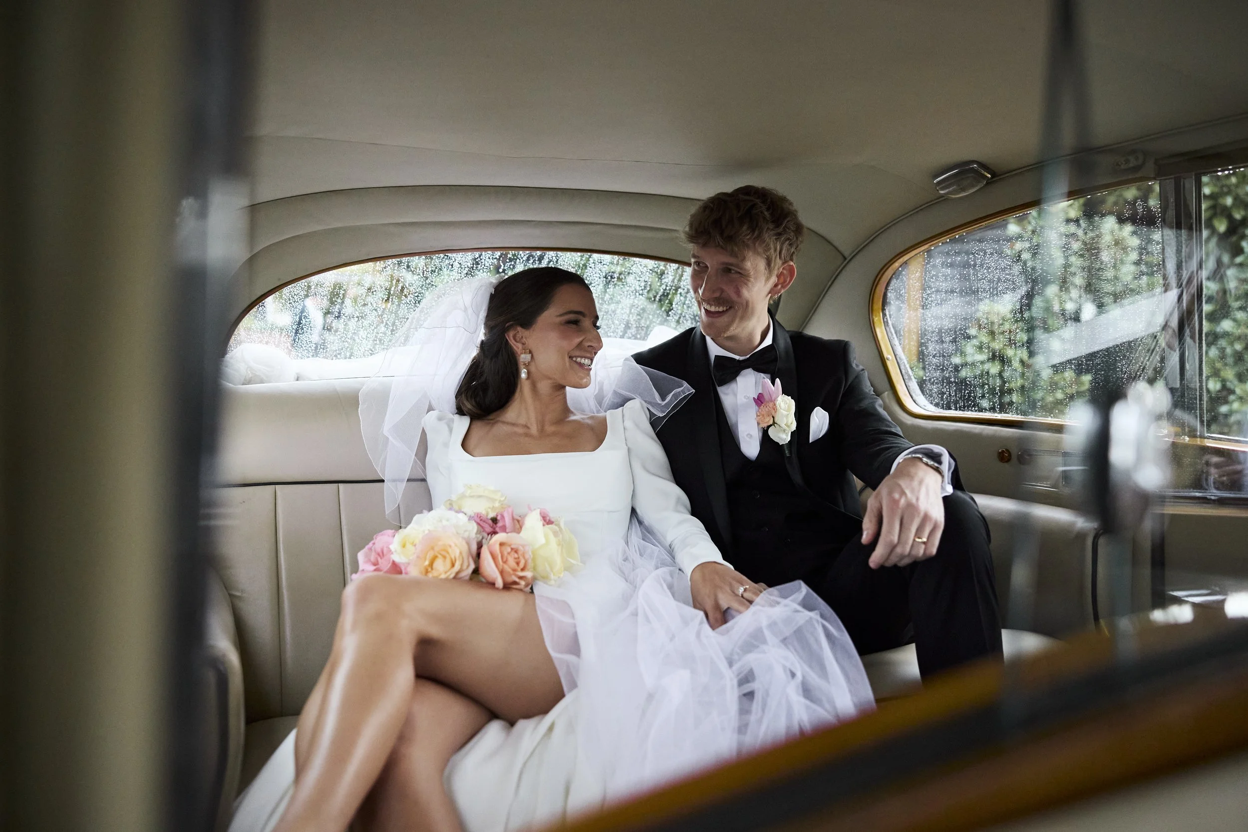 A bride and groom sitting inside a vintage car, smiling and looking at each other. The bride is holding a bouquet of flowers, and the groom is wearing a black tuxedo with a boutonniere. Rain is visible outside the car window.