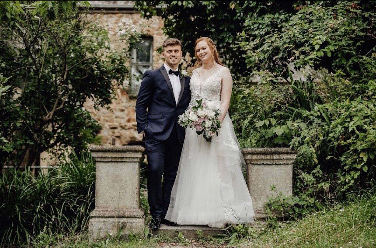 A bride and groom standing together outdoors, with lush greenery and stone walls in the background. The bride is holding a bouquet of flowers, and both are dressed in wedding attire.