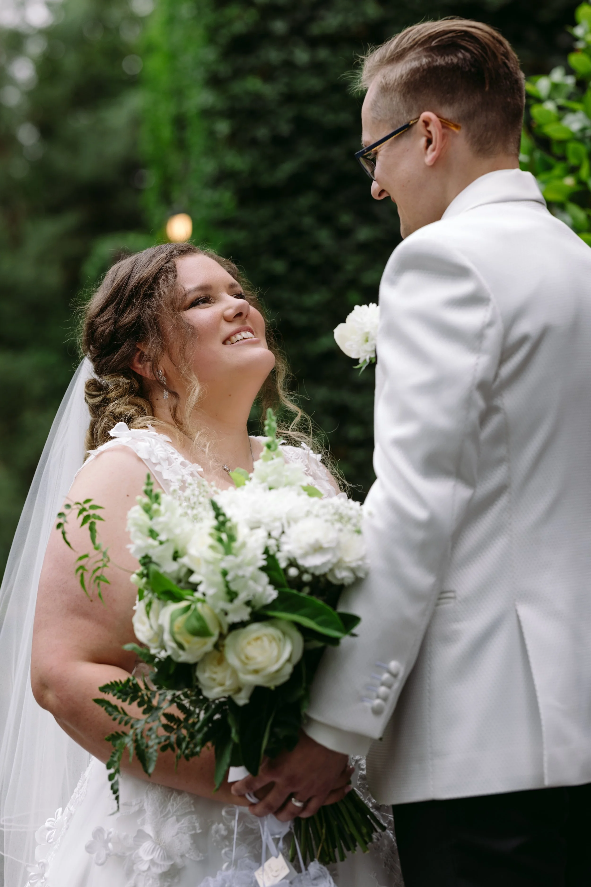 Bride and groom sharing a moment outdoors, with bride holding a bouquet of white flowers, trees in the background, and bride smiling up at groom.