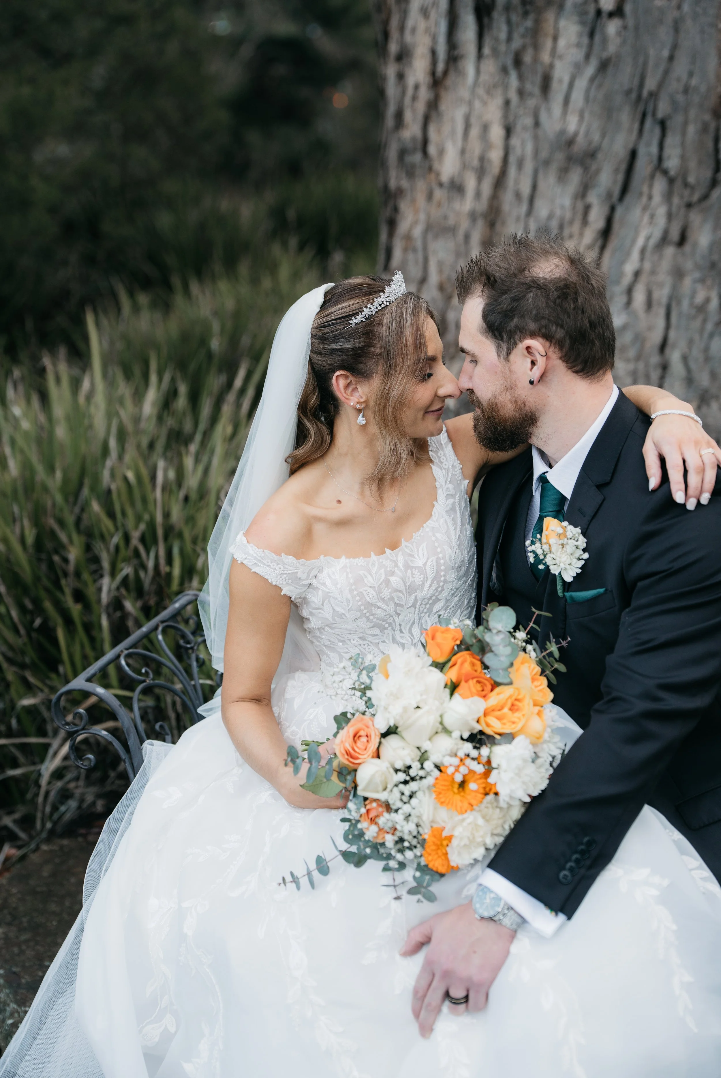 A bride and groom sitting close together on a bench, gazing into each other's eyes, outdoors near a large tree, with the bride holding a bouquet of orange, white, and greenery flowers.