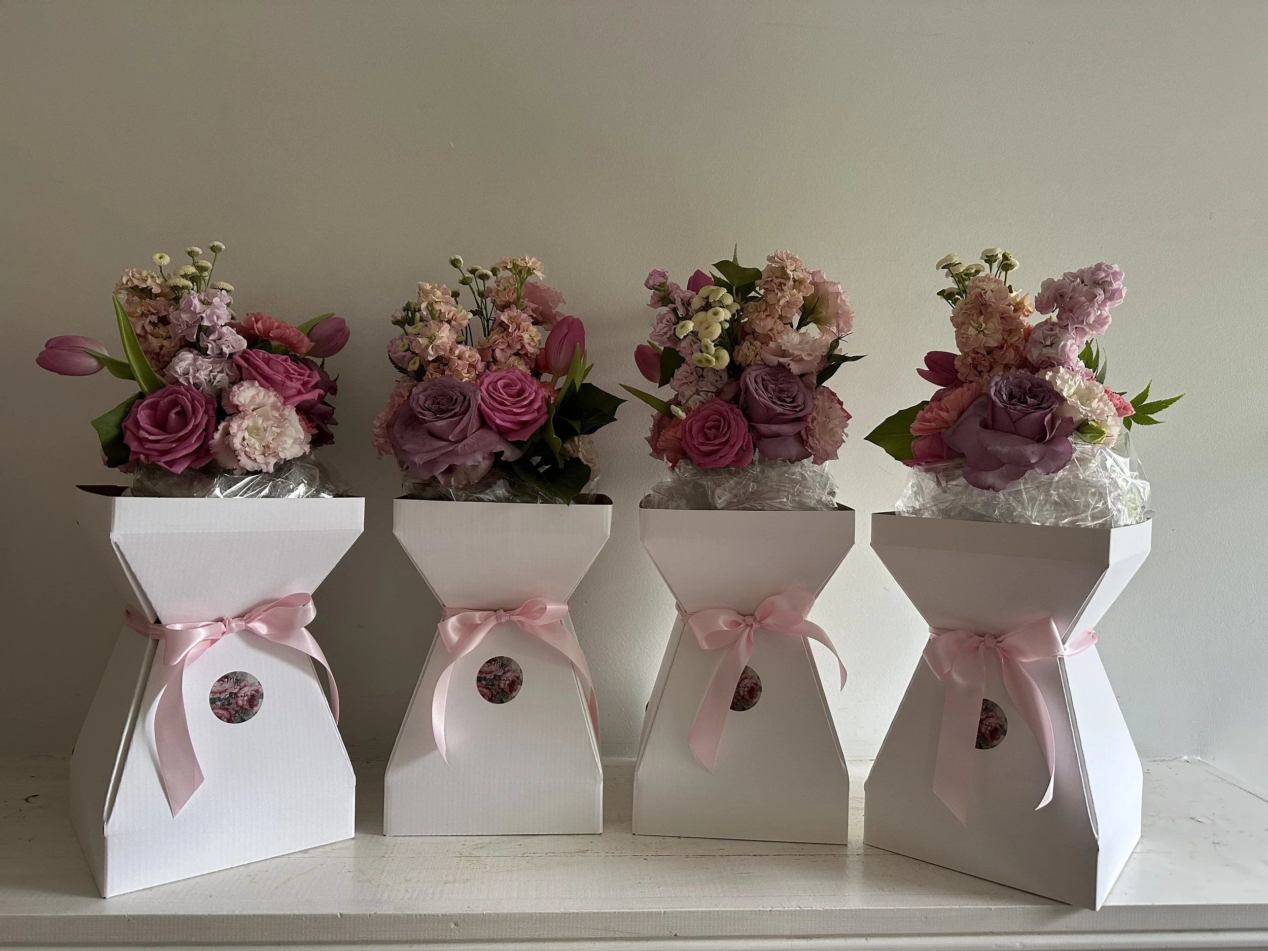 Four bouquets of pink and purple flowers in white gift boxes adorned with pink ribbons on a white surface against a plain wall.