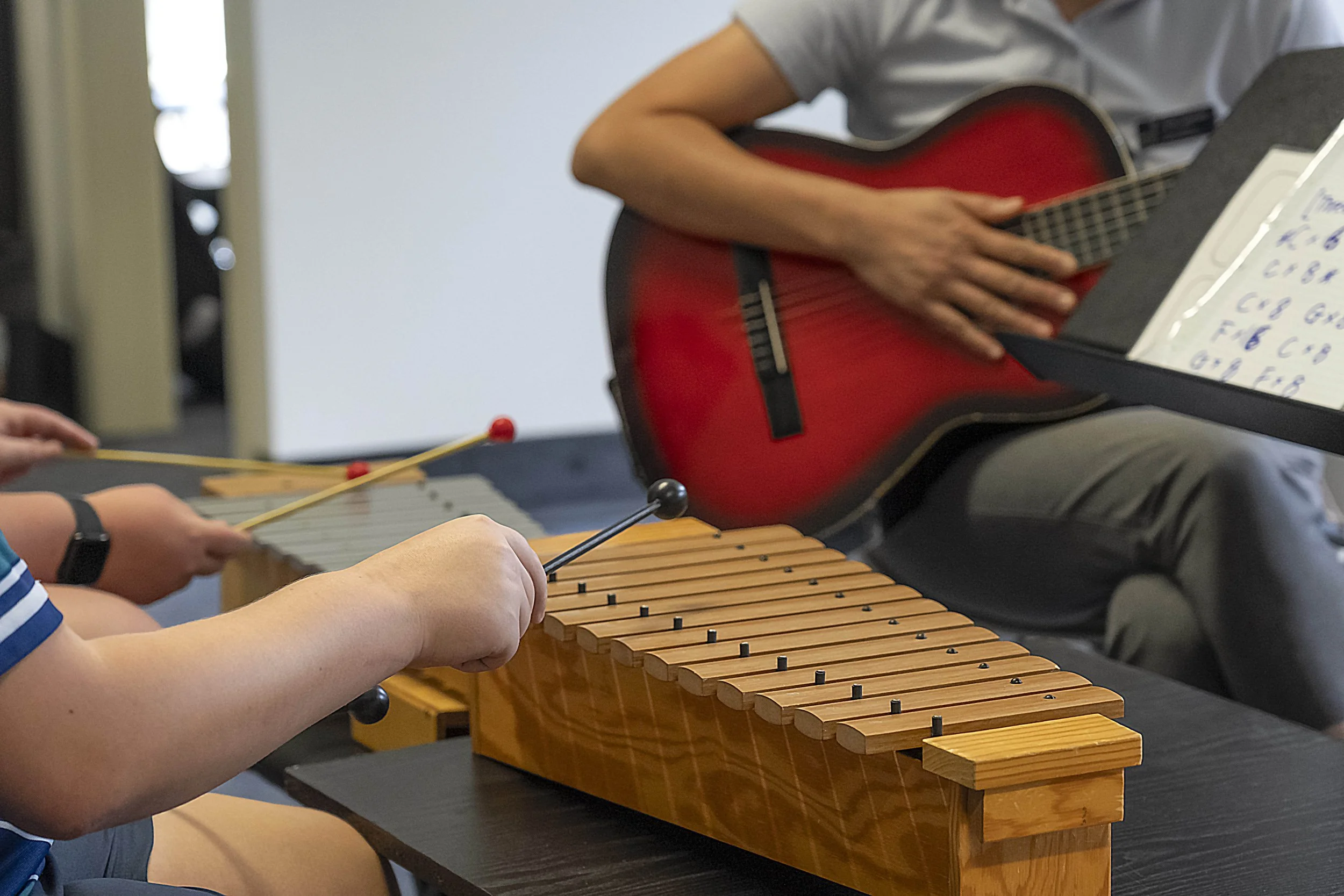 People playing musical instruments, including a marimba and a guitar, with sheet music in a music classroom.