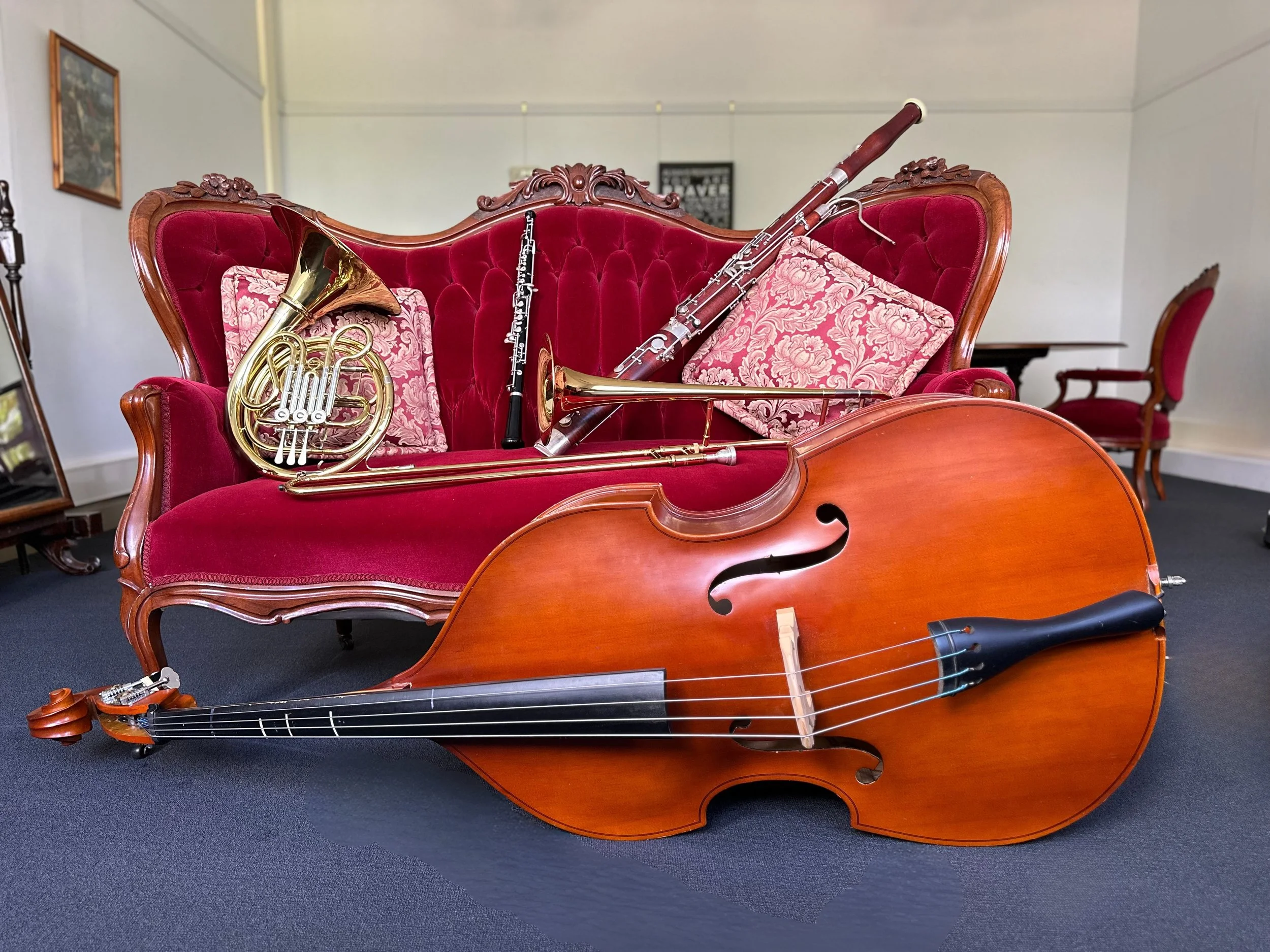 A vintage red velvet sofa with floral patterned pillows, a french horn, an oboe, a trombone, a bassoon and a large double bass on dark carpet in a room with white walls and a wooden table in the background.