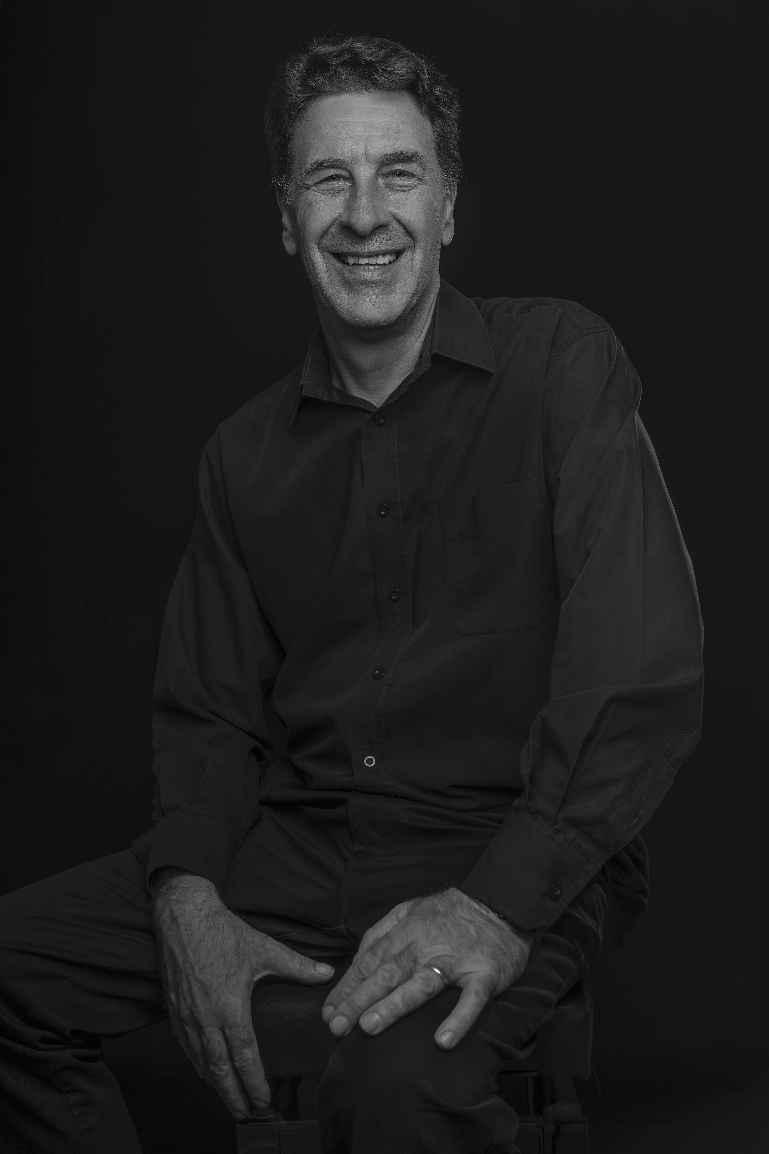 Black and white portrait of a smiling man with short hair, wearing a dark button-up shirt, sitting against a black background.