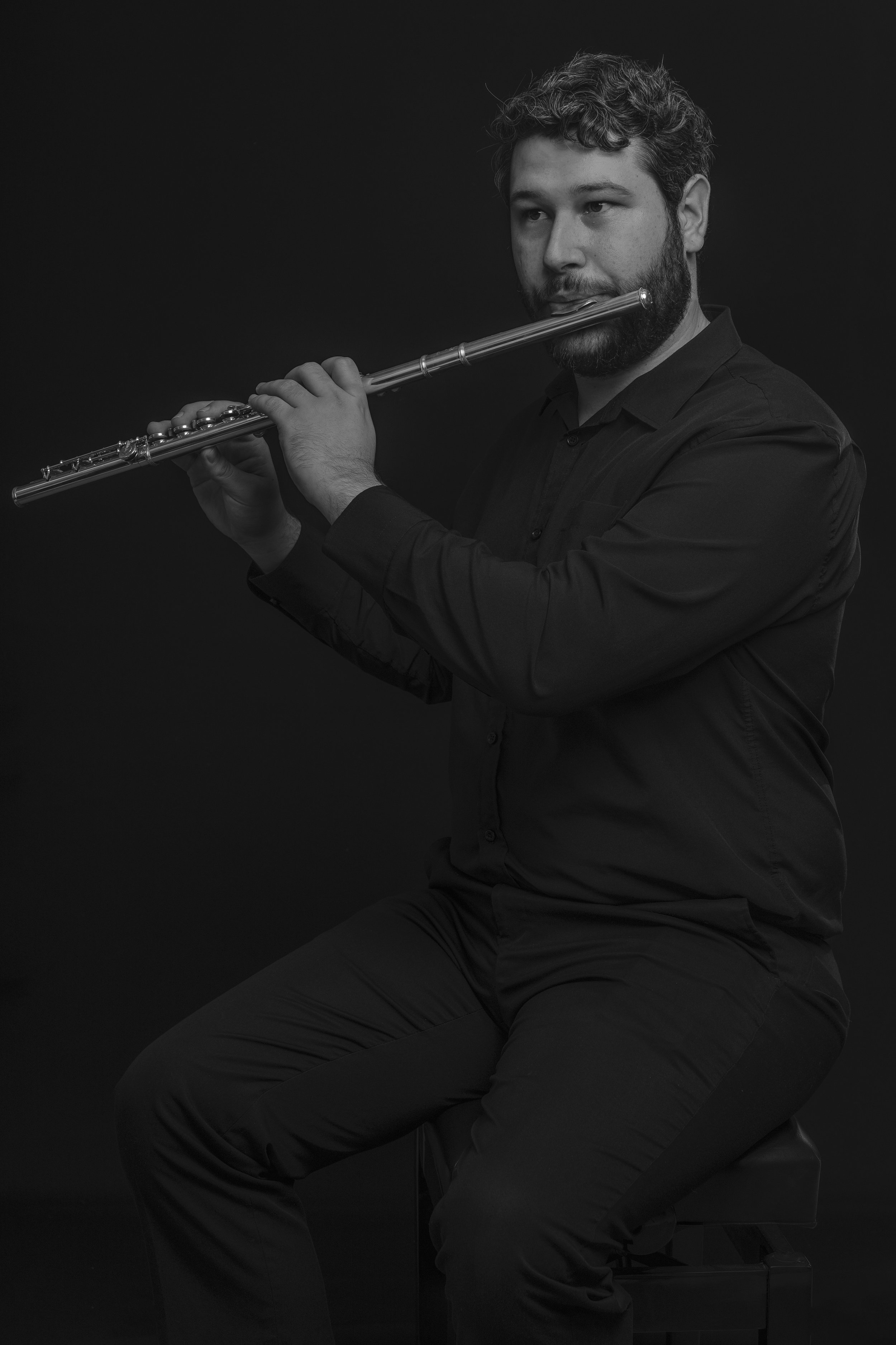 A man playing a flute, seated on a stool, dressed in black against a dark background.