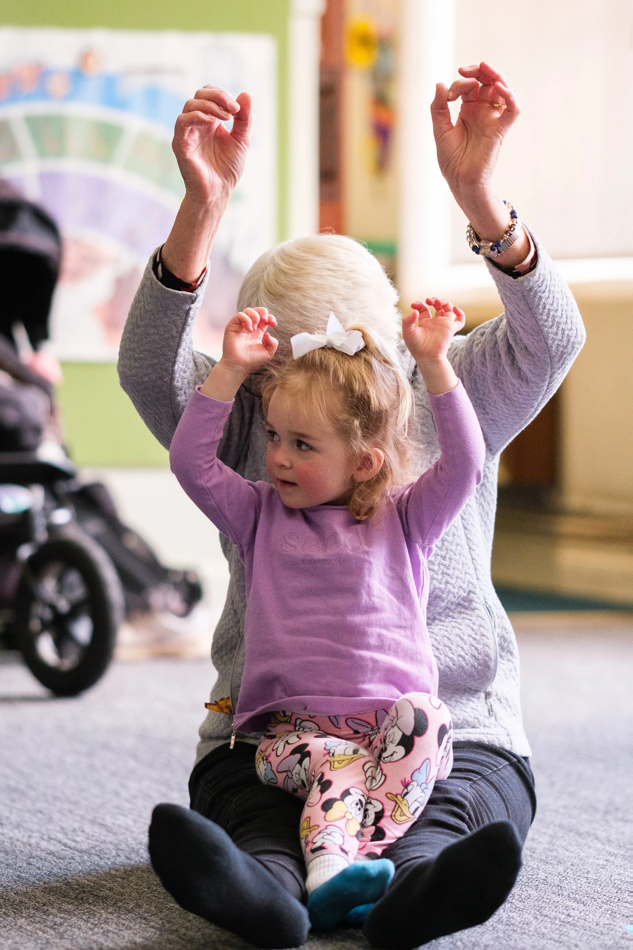 A young girl with curly hair, wearing pajamas with Disney characters, sitting on an adult's lap. The adult is behind her, raising hands, in a room with educational posters and a stroller nearby.