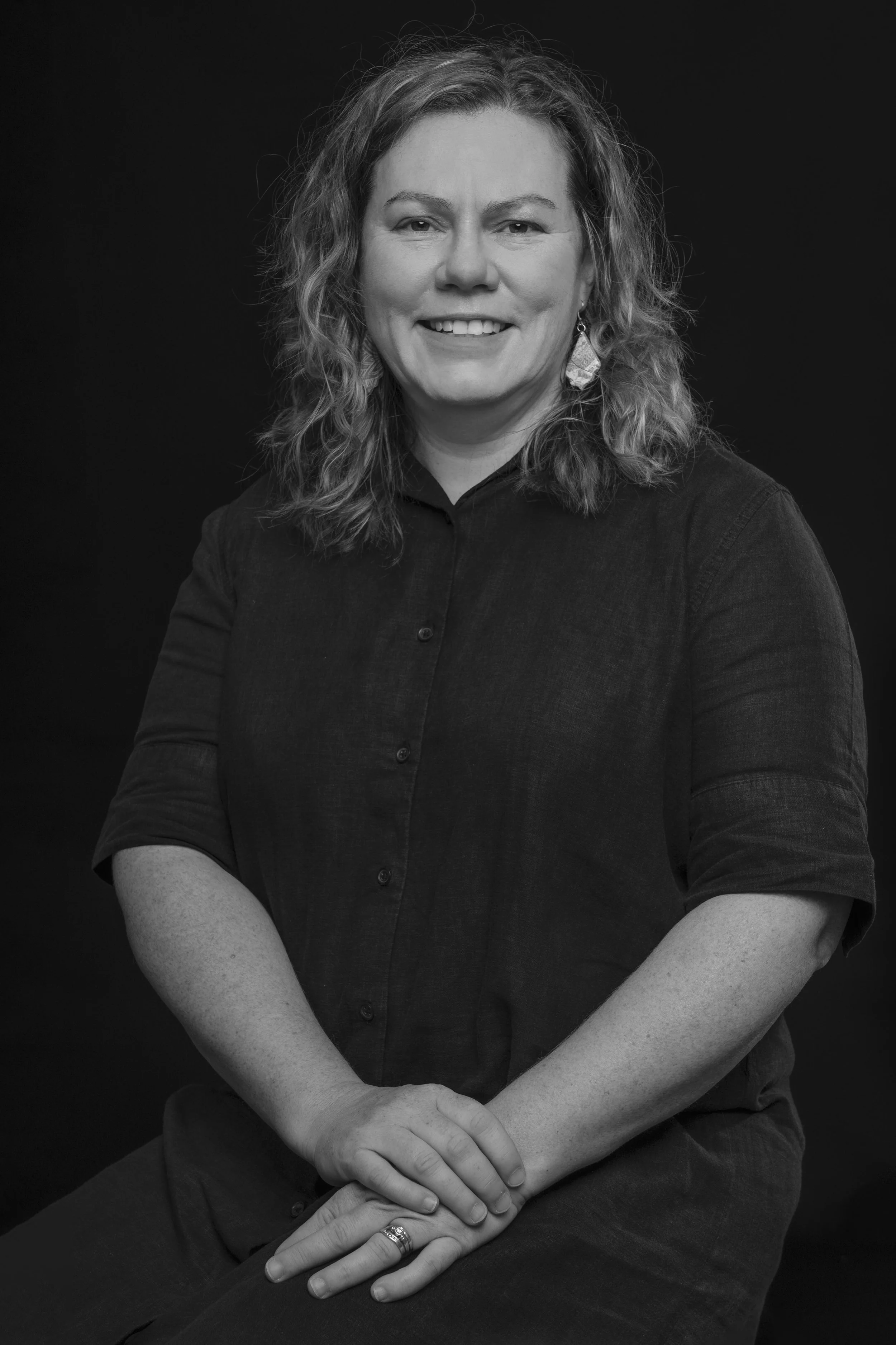 Black and white portrait of a woman with curly hair, smiling, wearing a dark button-up shirt and earrings, sitting against a dark background.