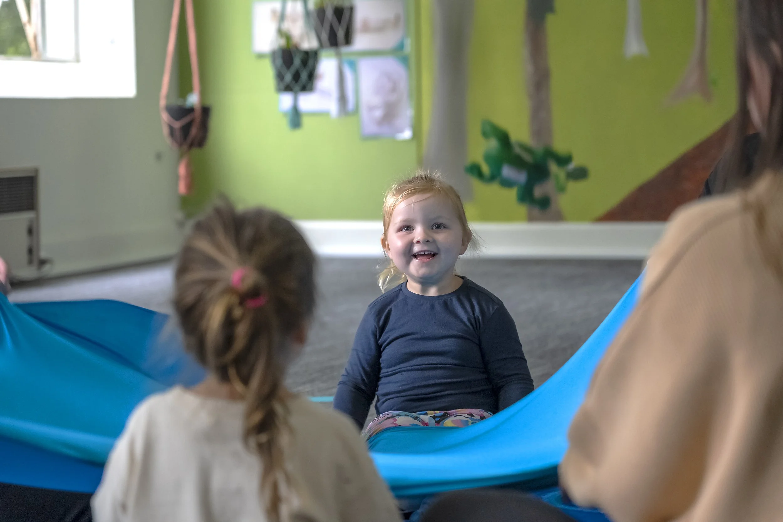 A young girl with red hair and a navy shirt smiling and sitting on a blue parachute in a room with green walls, while two other children sit nearby.