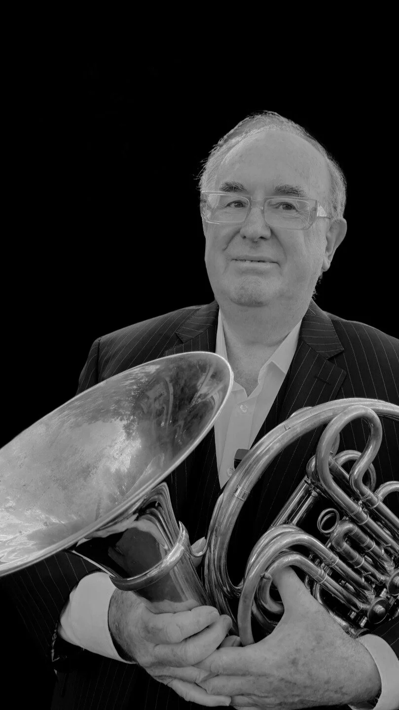 A man in a pinstripe suit holding a brass French horn, looking at the camera, with a black background.