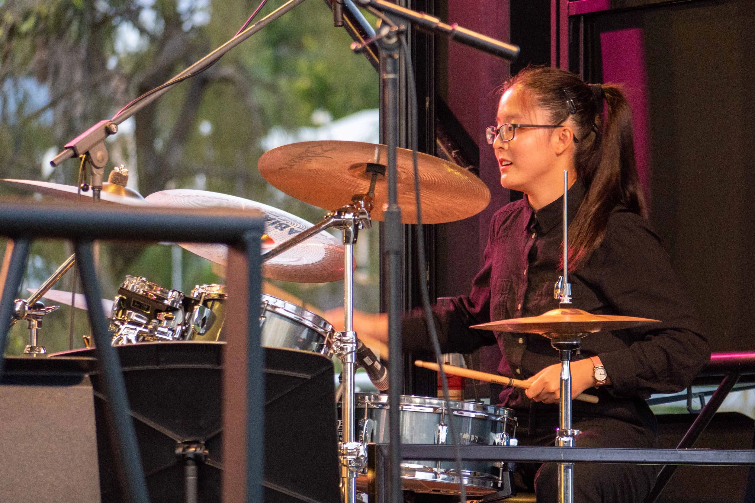 A young woman playing a drum set on stage during a performance, wearing glasses and a black shirt, with trees visible in the background.