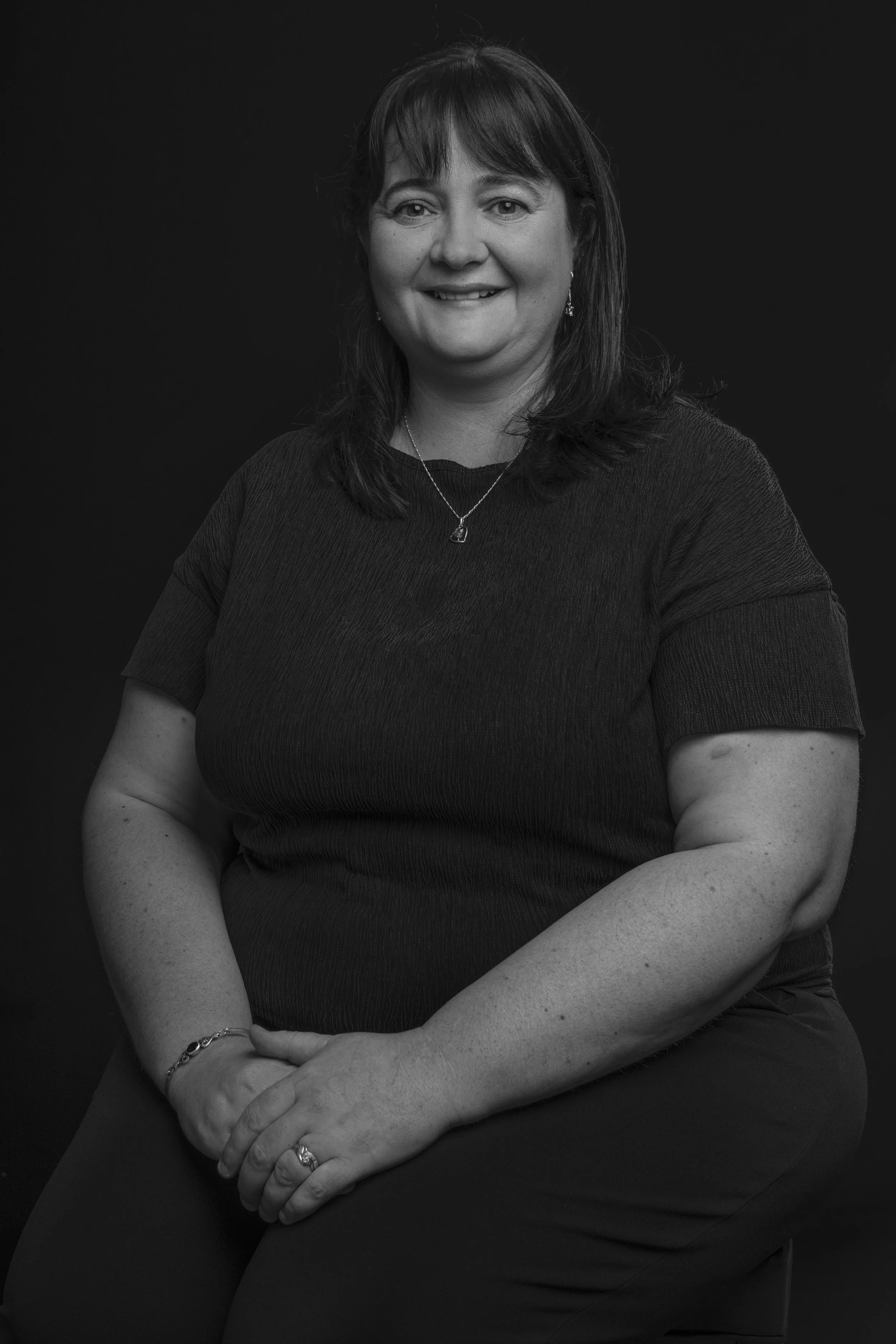 Black and white portrait of a woman with dark hair, smiling, wearing a short-sleeved top, necklace, bracelet, and ring, seated against a plain dark background.