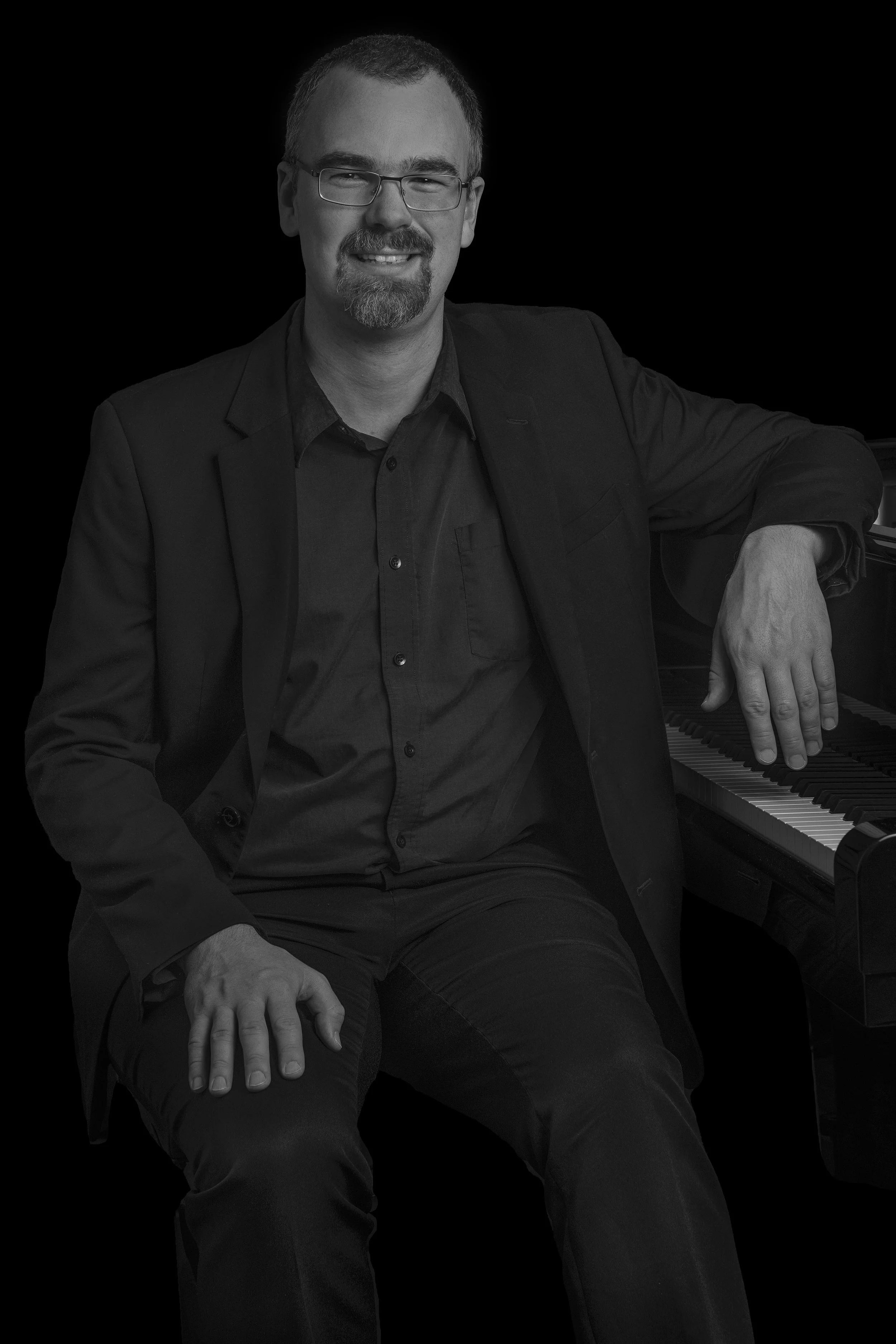 Man wearing glasses and a dark suit sitting next to a piano, smiling at the camera.