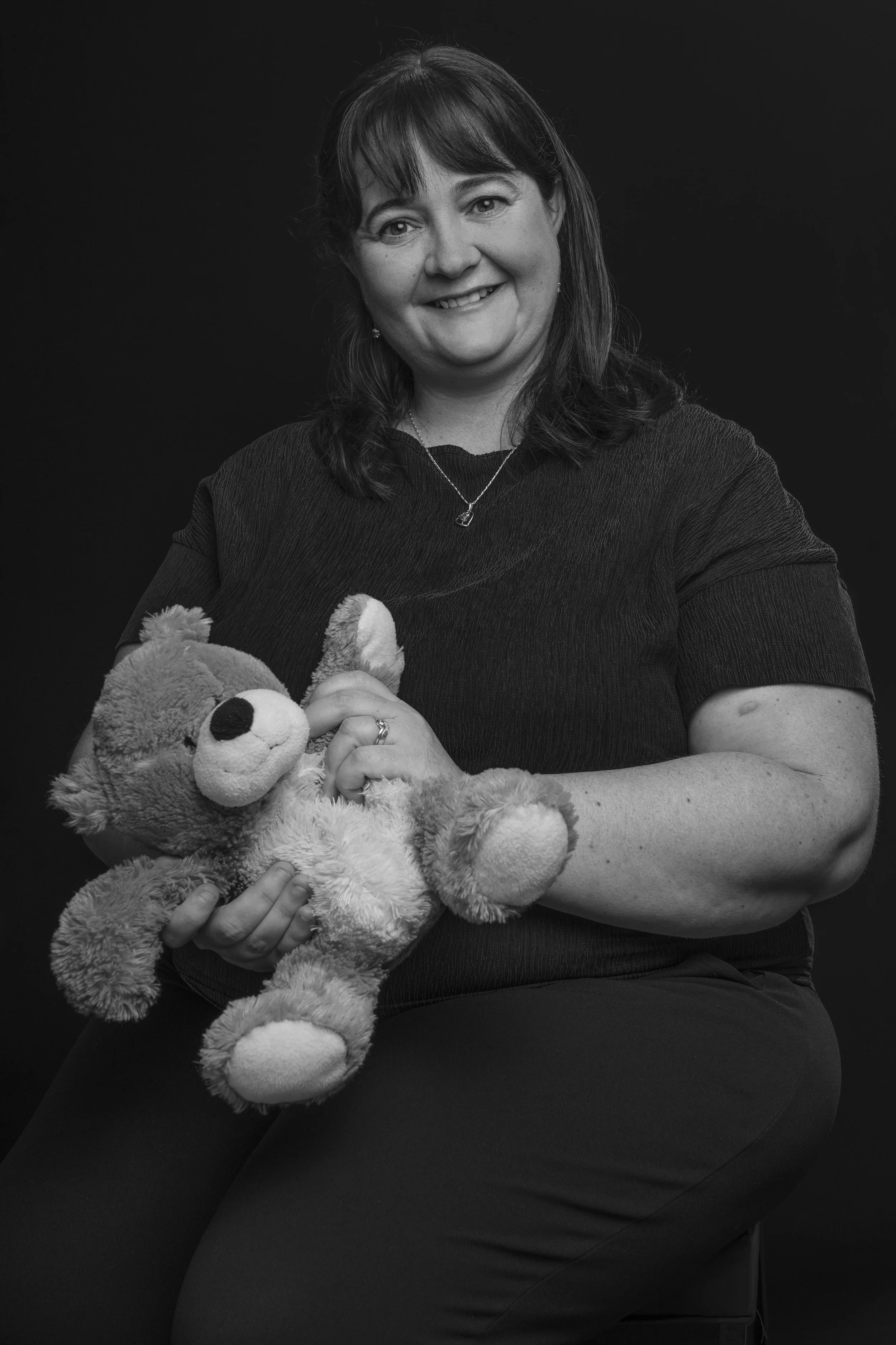 A woman smiling while holding a teddy bear, sitting against a dark background.