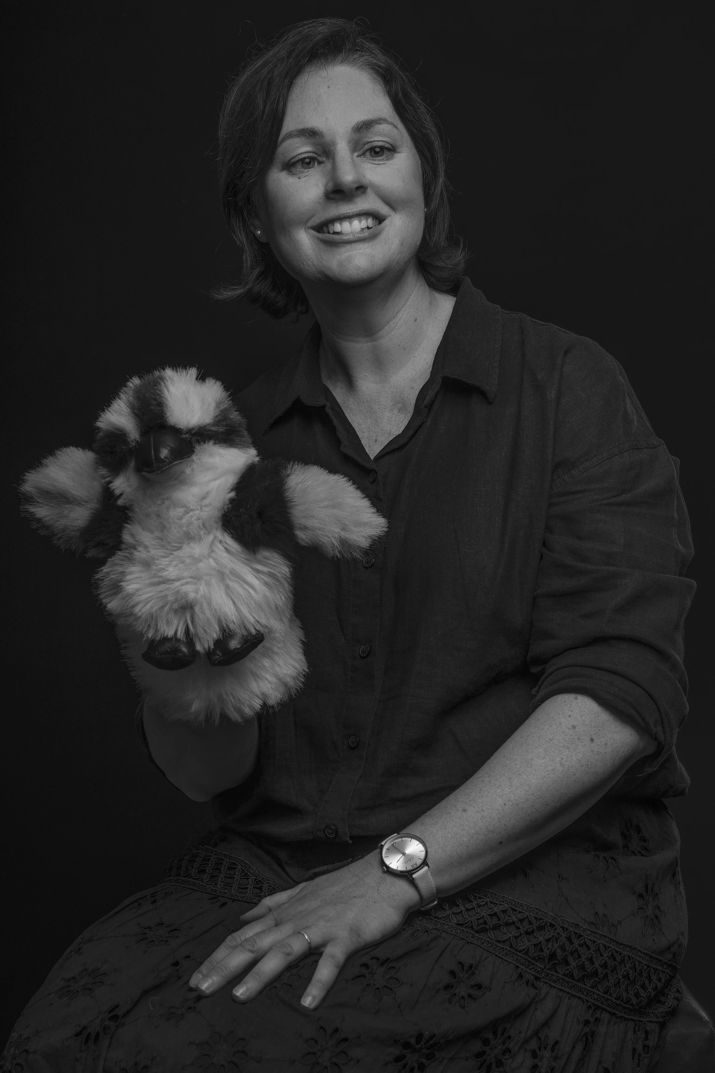 A woman with shoulder-length hair smiling, sitting against a dark background, holding a plush stuffed kookaburra.