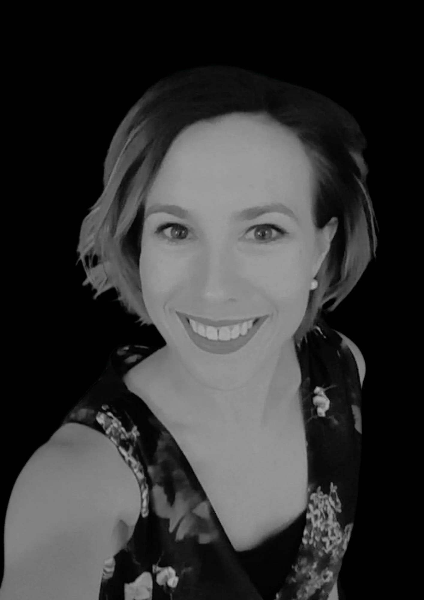 Black and white portrait of a woman smiling, with shoulder-length hair, wearing a floral sleeveless top, against a dark background.