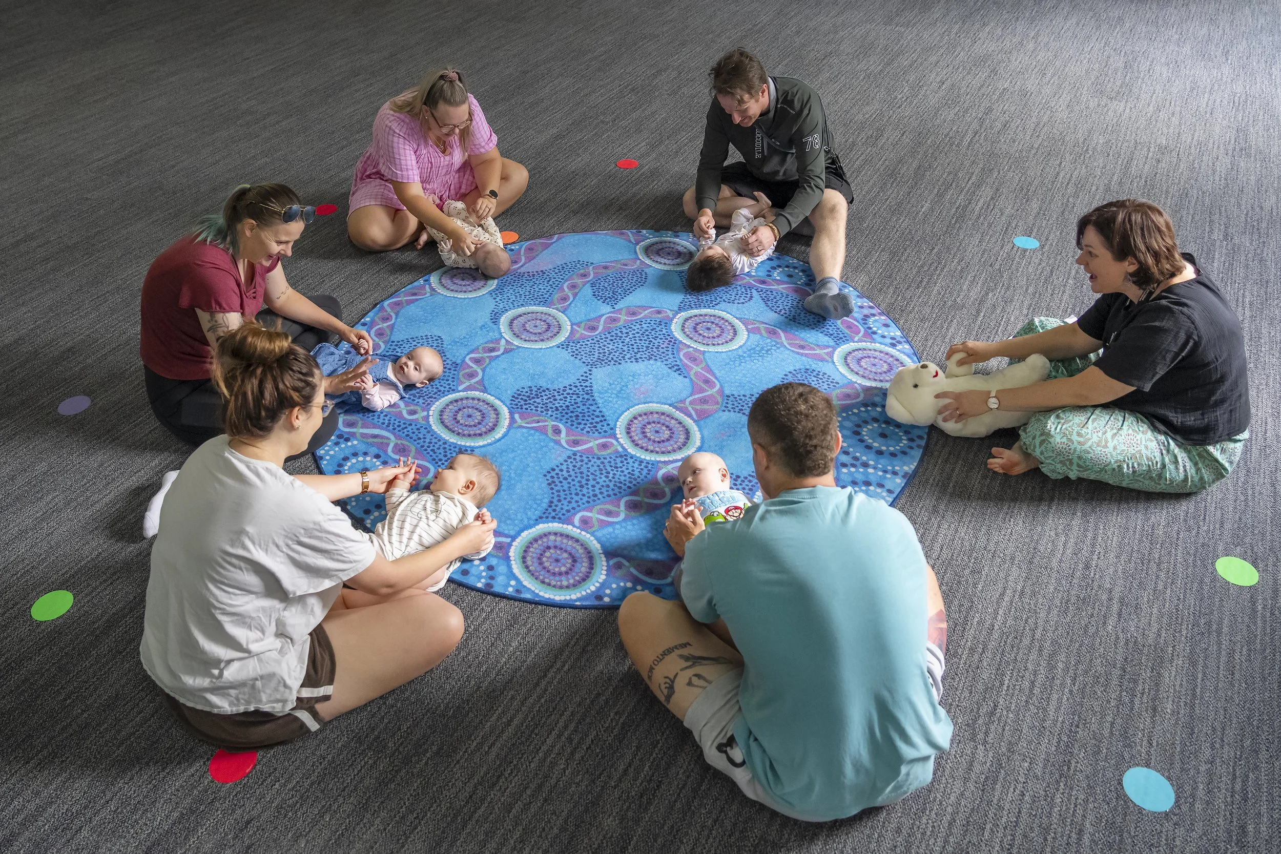Group of seven adults and four babies sitting on a circular blue patterned rug, engaging in a baby sensory class. The adults are holding and playing with the babies, some using toys or cuddling them. The room has gray carpet with colorful dots scattered around.