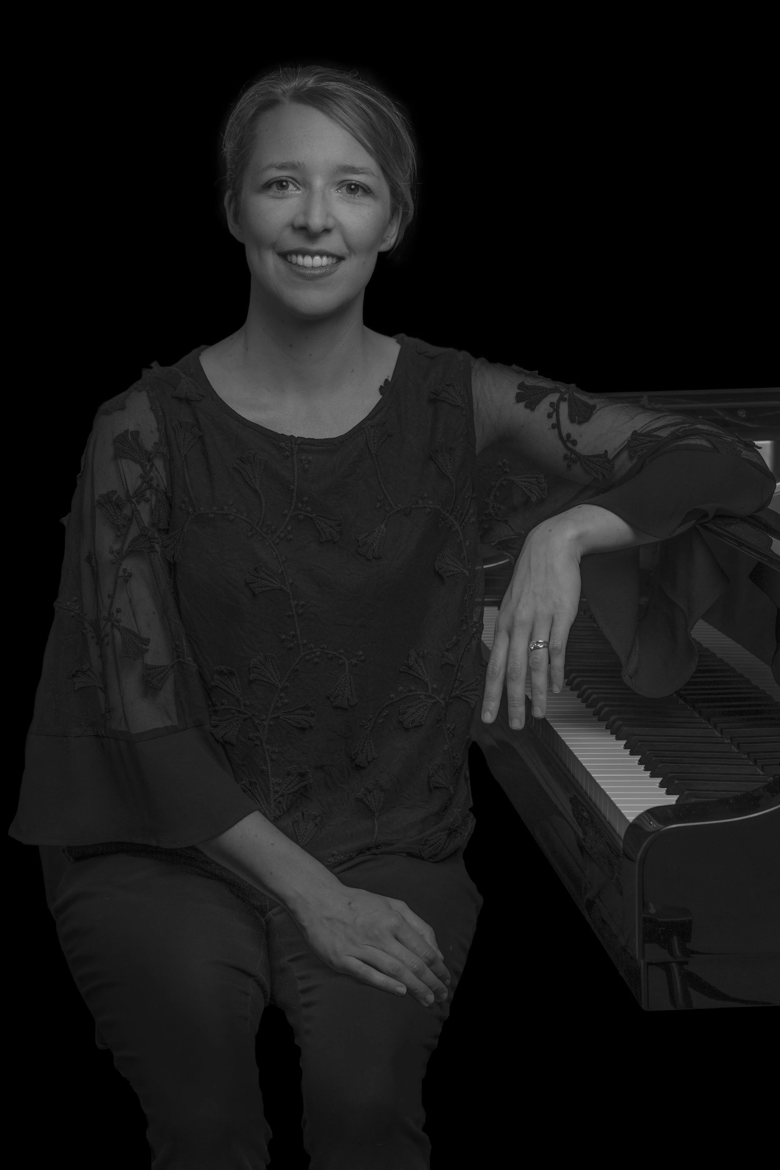 A woman sitting next to a grand piano, smiling, wearing a black embroidered blouse, with her hand resting on the keyboard.