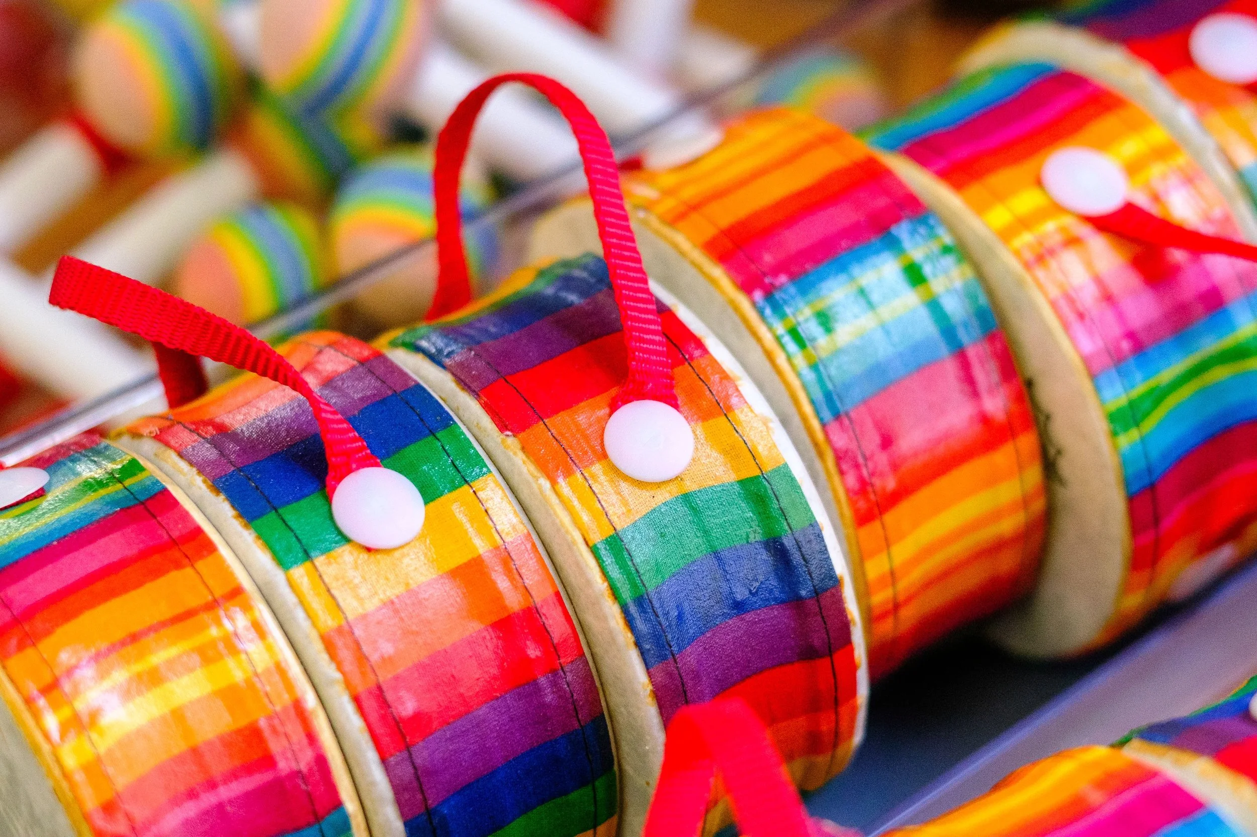 Rainbow drums with rainbow stripe pattern hanging on pins, with a red ribbon looped through each, displayed in a row.