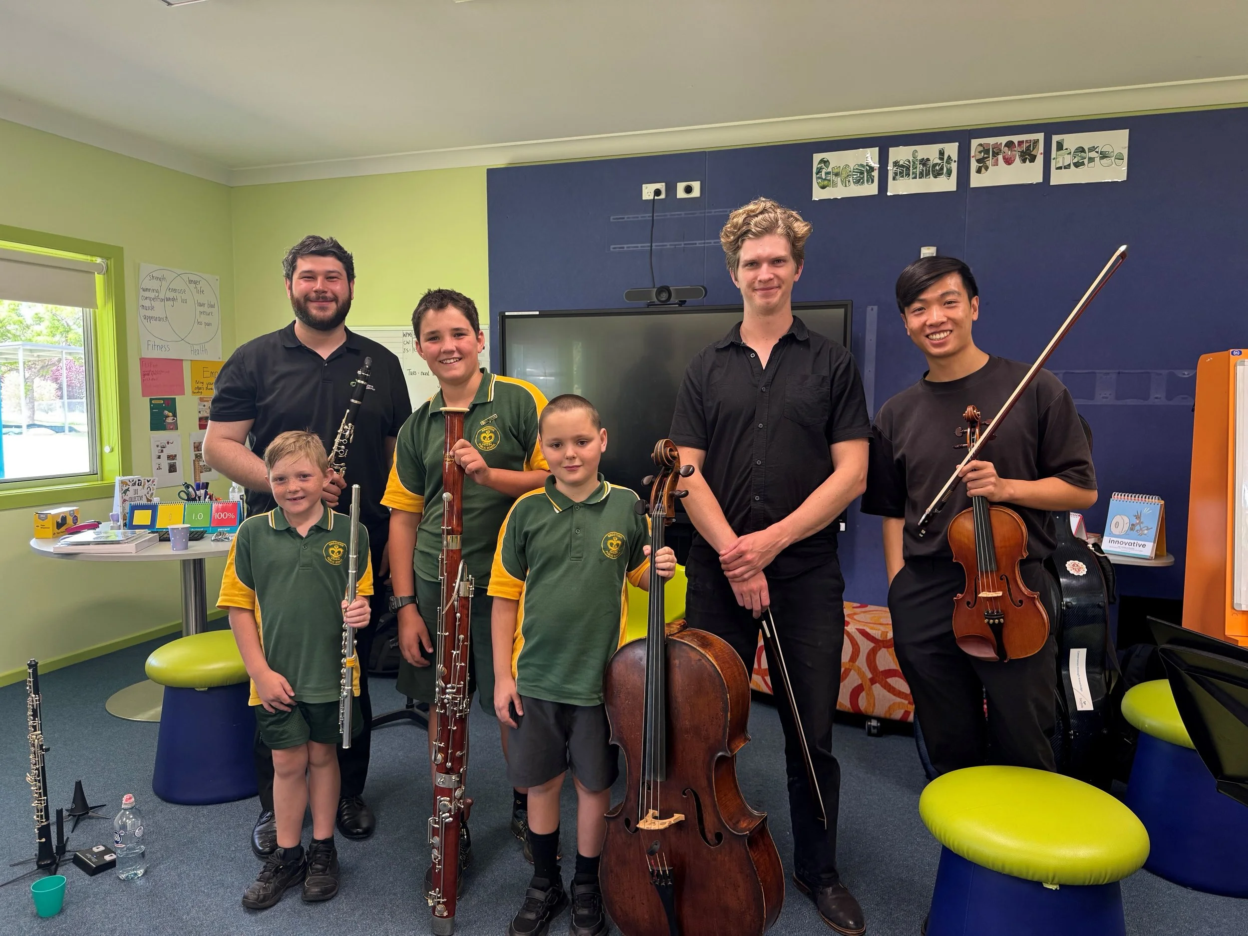 Group of five children and two adults standing in a classroom with musical instruments, including a violin, cello, flute, clarinet, and bassoon.