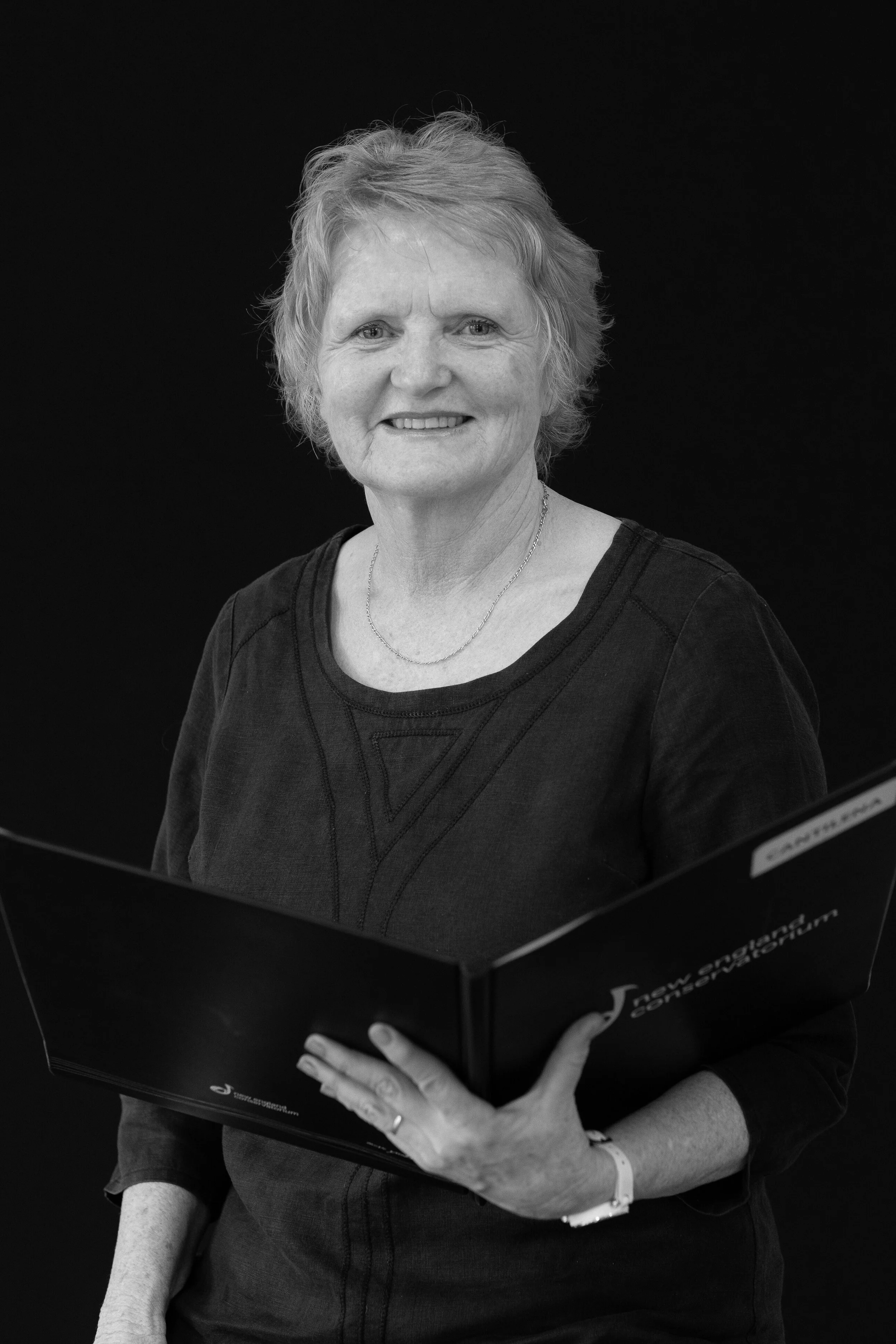 A woman with short, wavy hair, wearing a dark top and a necklace, smiling while holding a choral folder, against a solid dark background.
