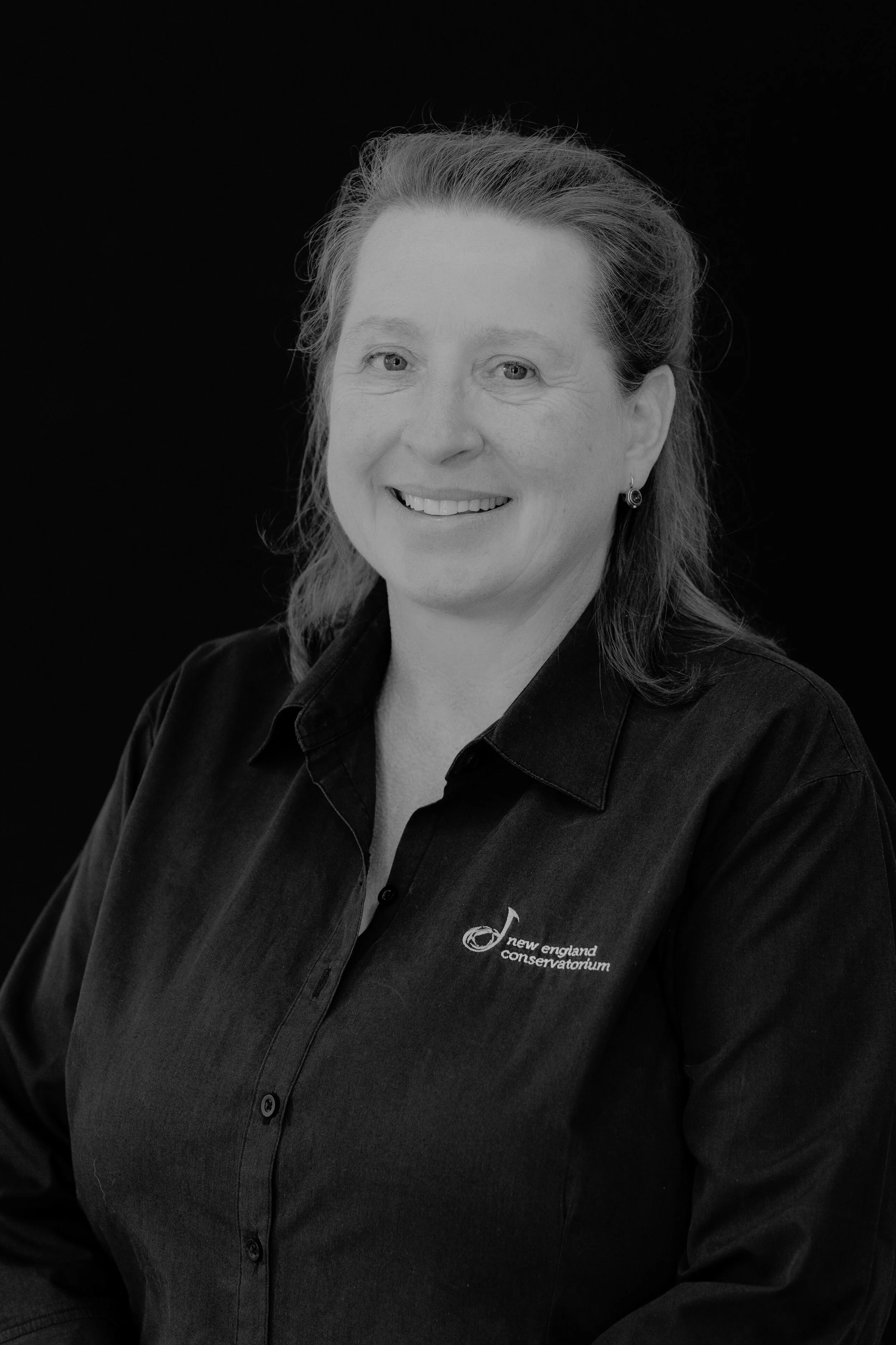 Black and white portrait of a woman smiling, wearing a dark shirt with a logo that says New England Conservatorium against a dark background.