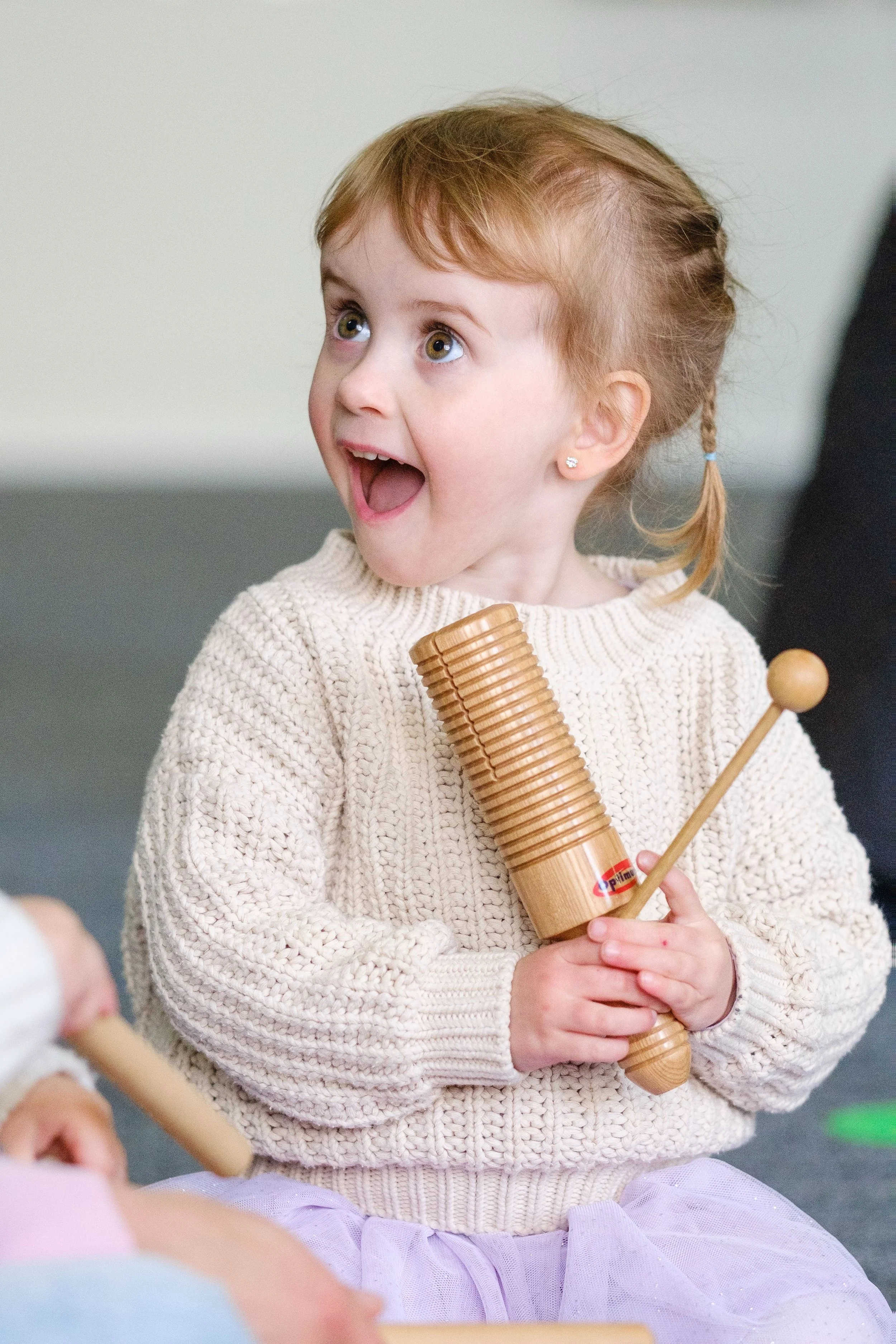 Smiling young girl with blonde hair in a braid, wearing a cream sweater, holding a wooden percussion instrument and a mallet, indoor setting.