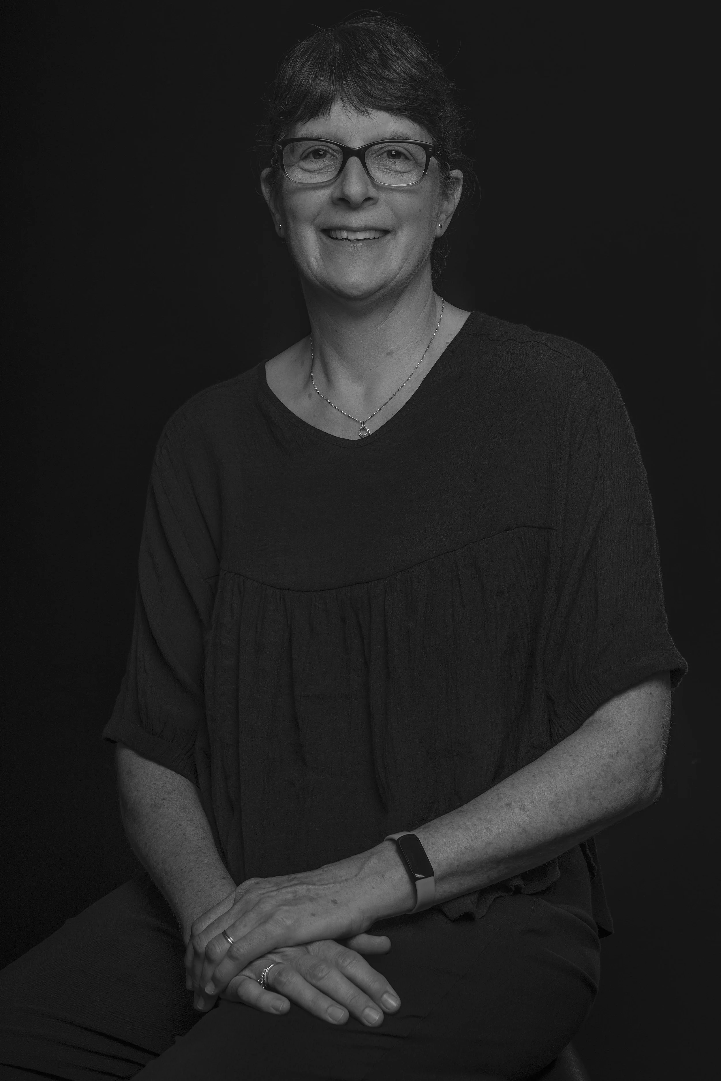 Black and white portrait of a smiling woman wearing glasses, a necklace, and a dark blouse, sitting against a dark background.