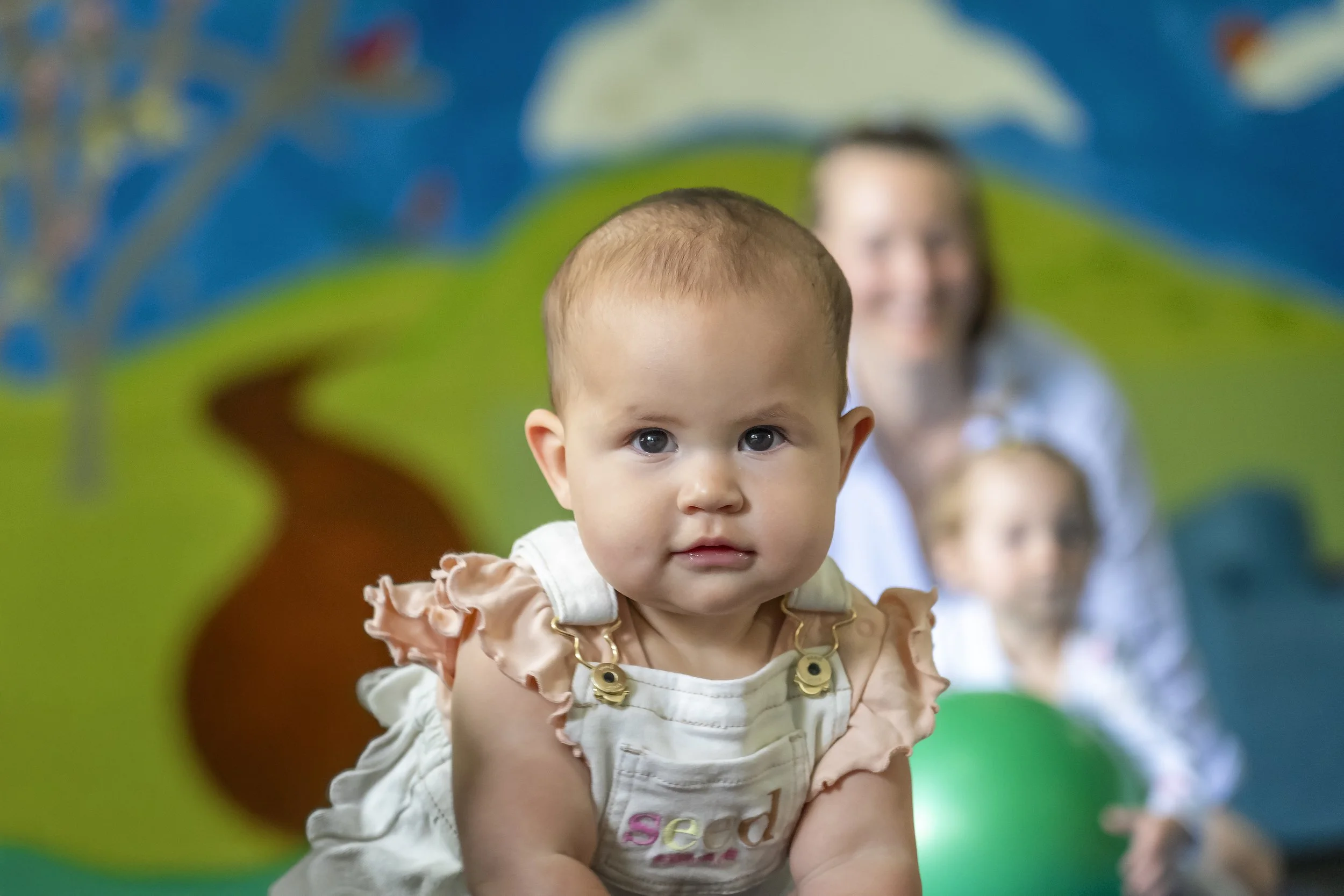 A baby girl crawling on a colorful play area with a painted mural background, with a woman and another child blurred behind her.