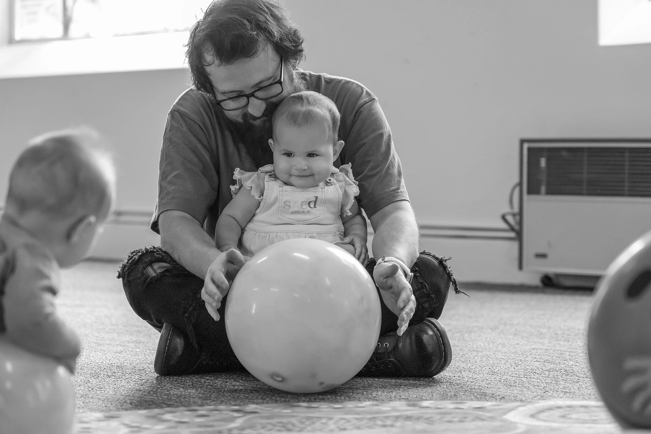 A man with glasses and a beard is sitting on the floor with a young girl on his lap, both looking at a balloon. The girl is smiling and wearing a dress that says "Seed." There are children nearby in a room with a carpeted floor and a window.