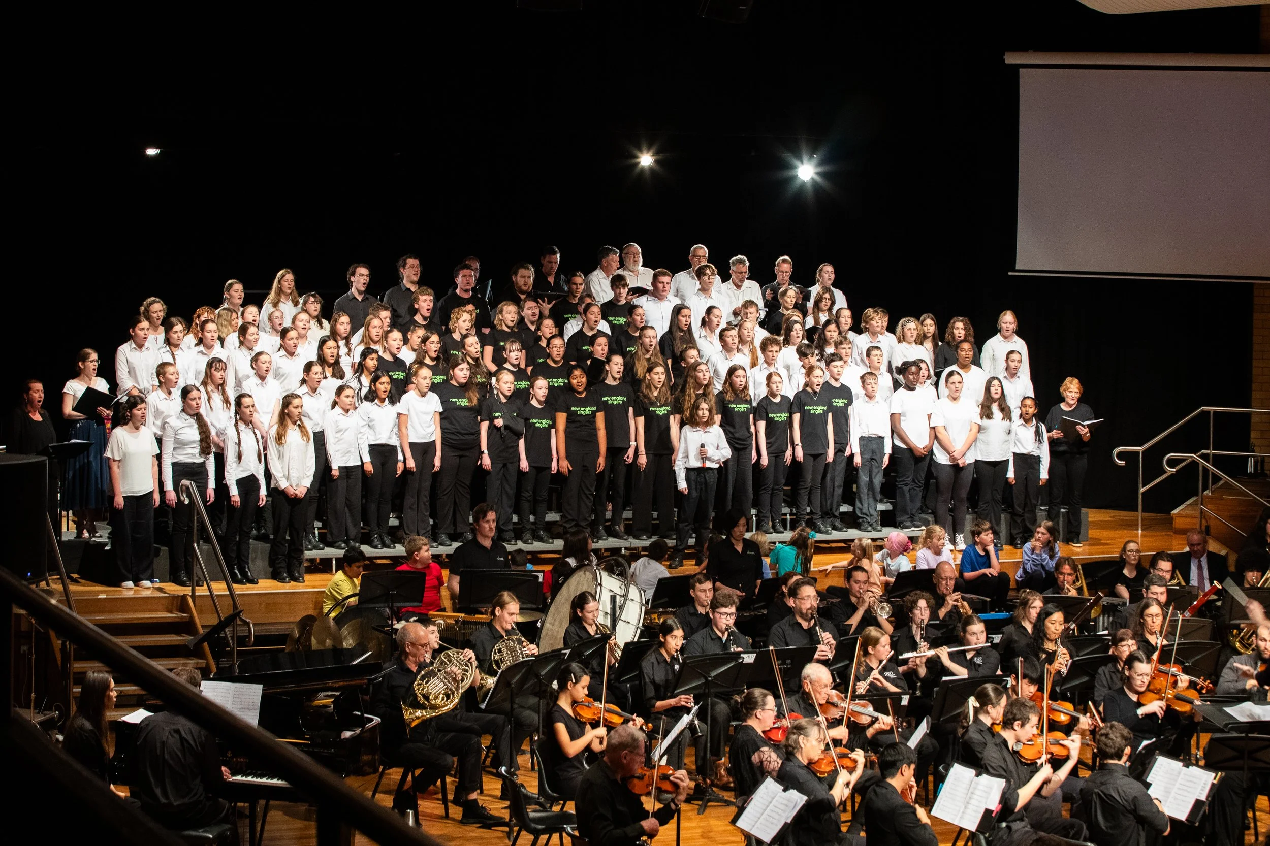 A large mixed-voice choir singing on stage, accompanied by an orchestra, during a concert. The choir members are arranged in multiple rows, wearing black and white attire. The orchestra includes string, brass, and percussion instruments, with musicians seated in front of the choir.