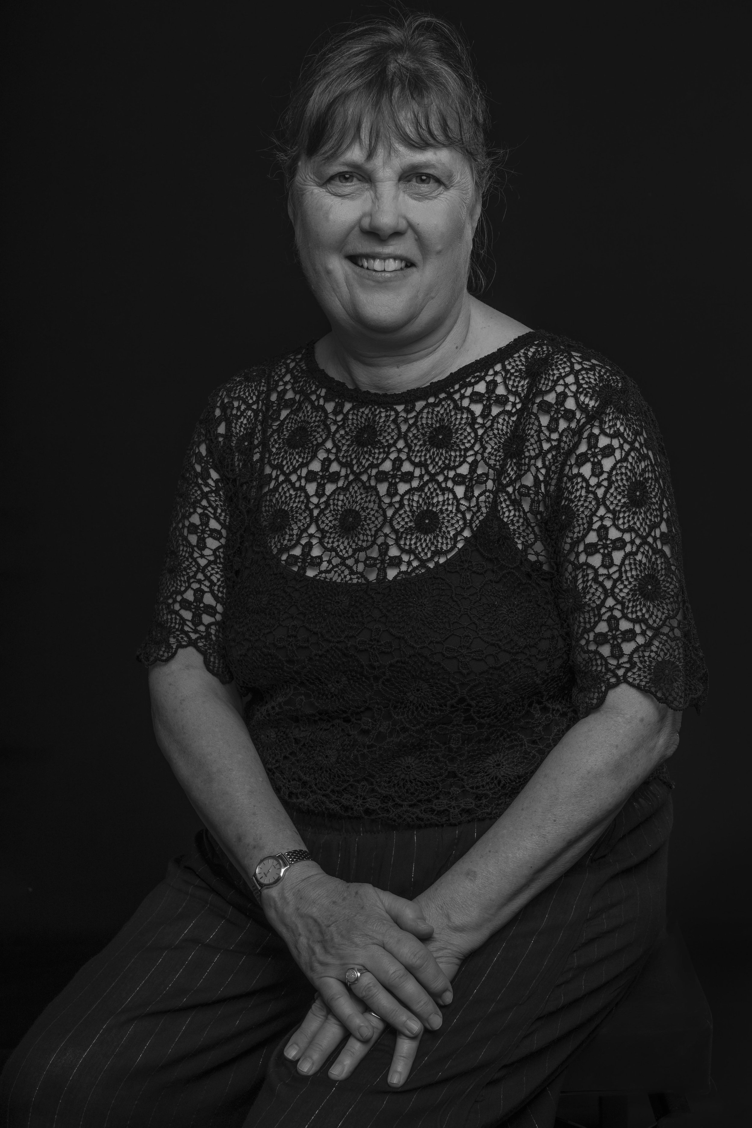 Black and white portrait of a smiling woman with short hair, wearing a lace top and striped pants, seated with hands resting on her lap against a dark background.