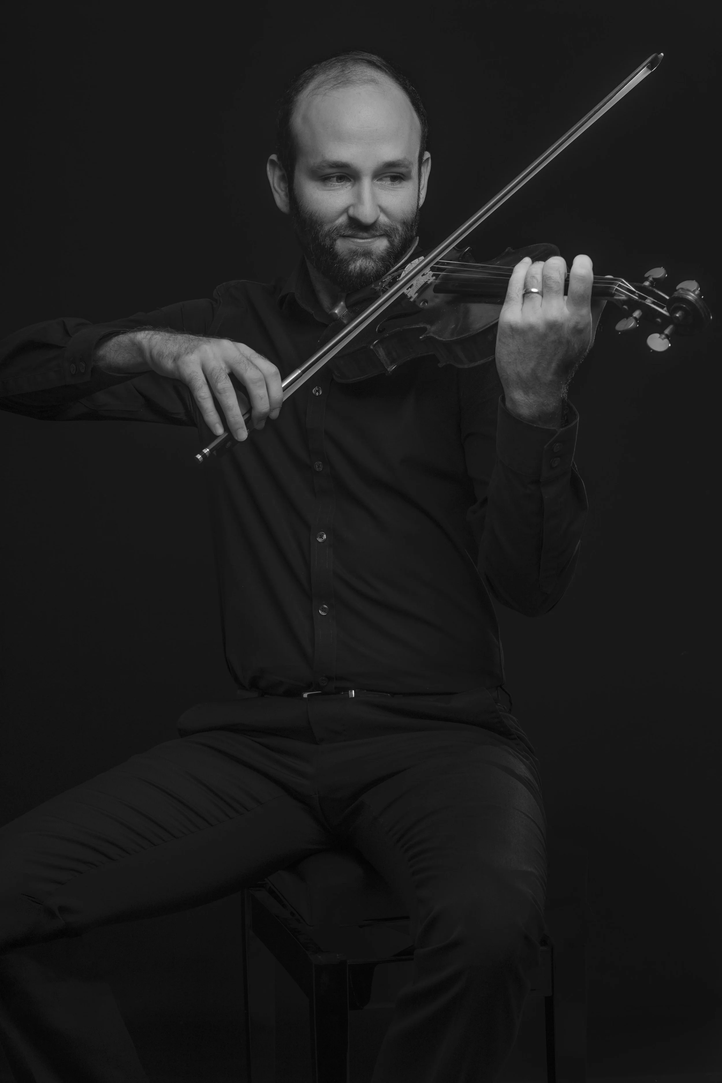 A man with a beard playing the violin, seated against a dark background, wearing a black shirt and appearing to enjoy the music.