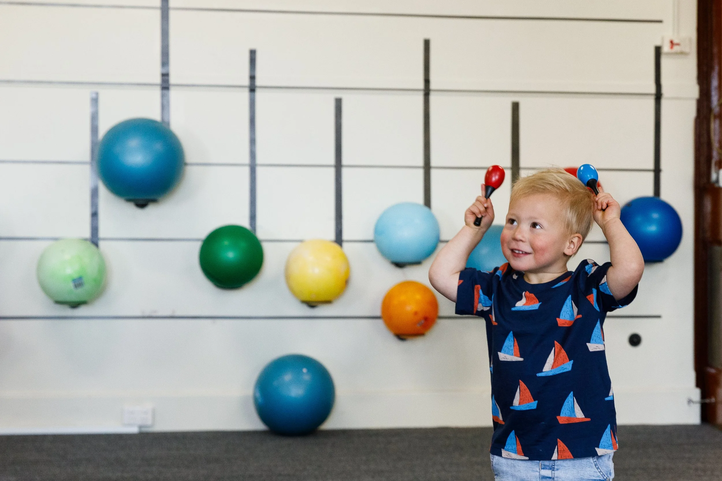A young boy wearing a blue shirt with sailboats, holding maracas, standing in front of a wall with colored exercise balls.