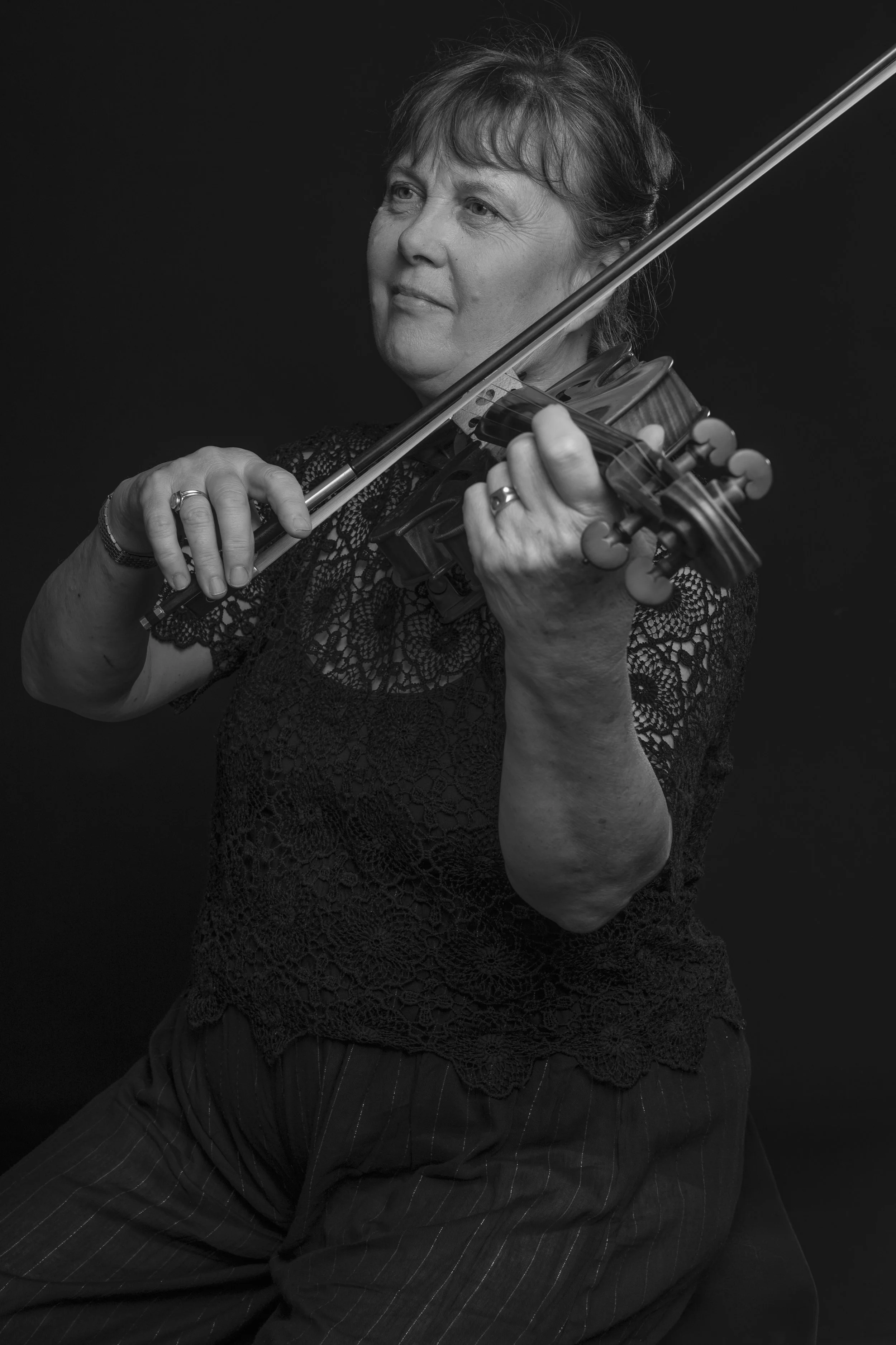 Black and white photo of a woman playing the violin, wearing a lace top and striped pants, against a dark background.
