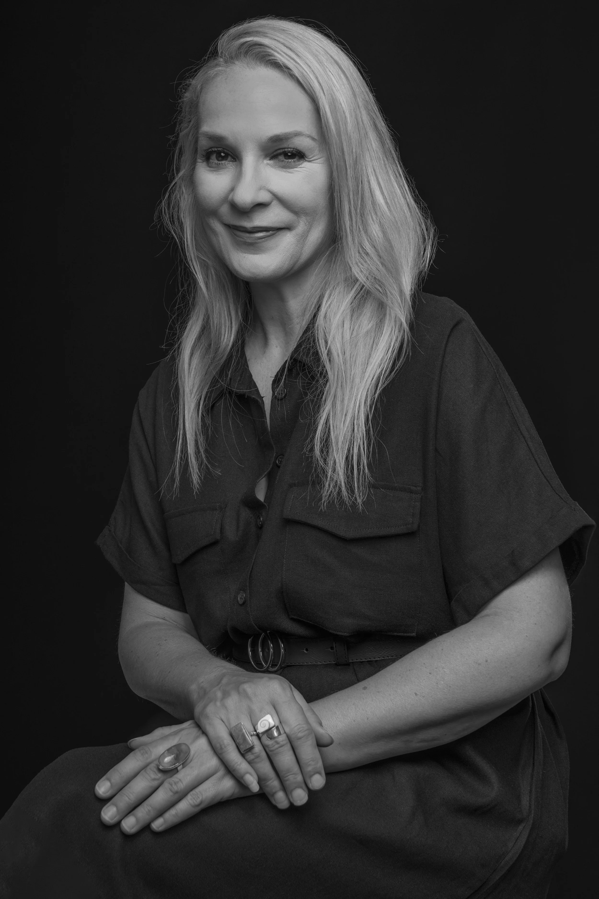 Black and white portrait of a woman with long blonde hair, smiling slightly, wearing a short-sleeved button-up shirt with two chest pockets, sitting with her hands resting on her lap, and wearing rings and a belt.