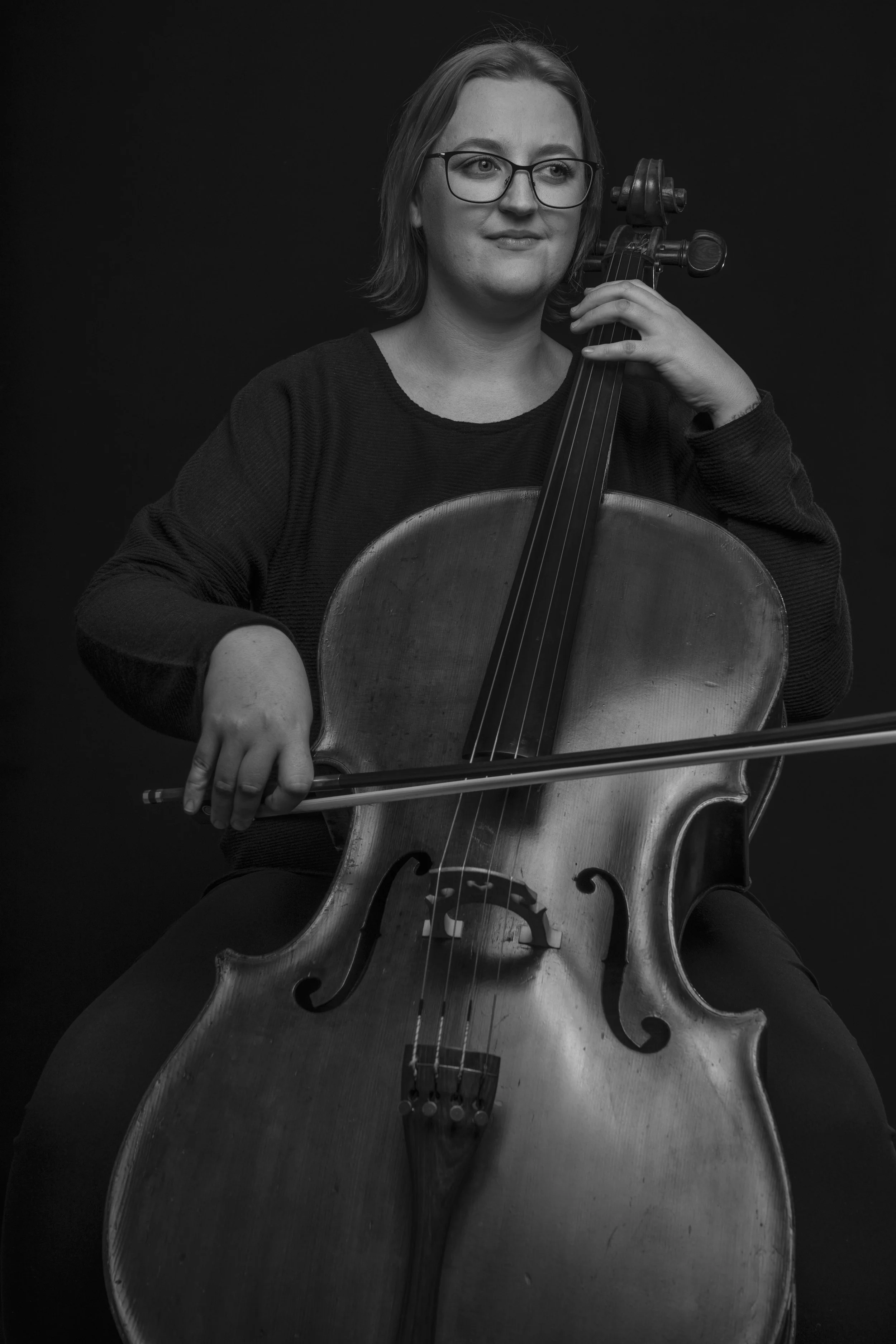 A woman with glasses is sitting and holding a cello and bow, looking slightly to the side, in a black and white photo.
