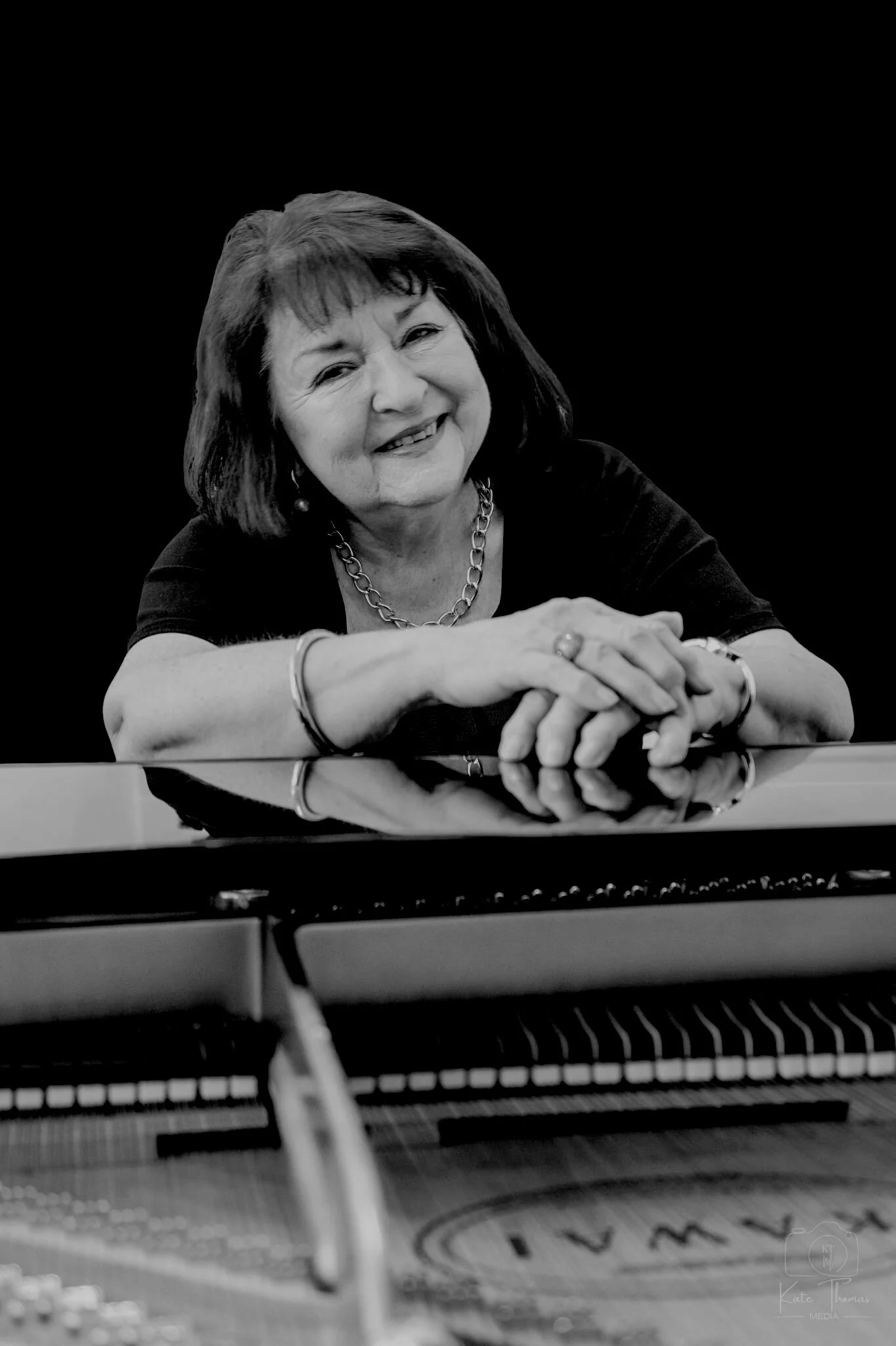 Black and white photo of a smiling woman with shoulder-length hair, sitting at a grand piano with her arms resting on it, against a dark background.