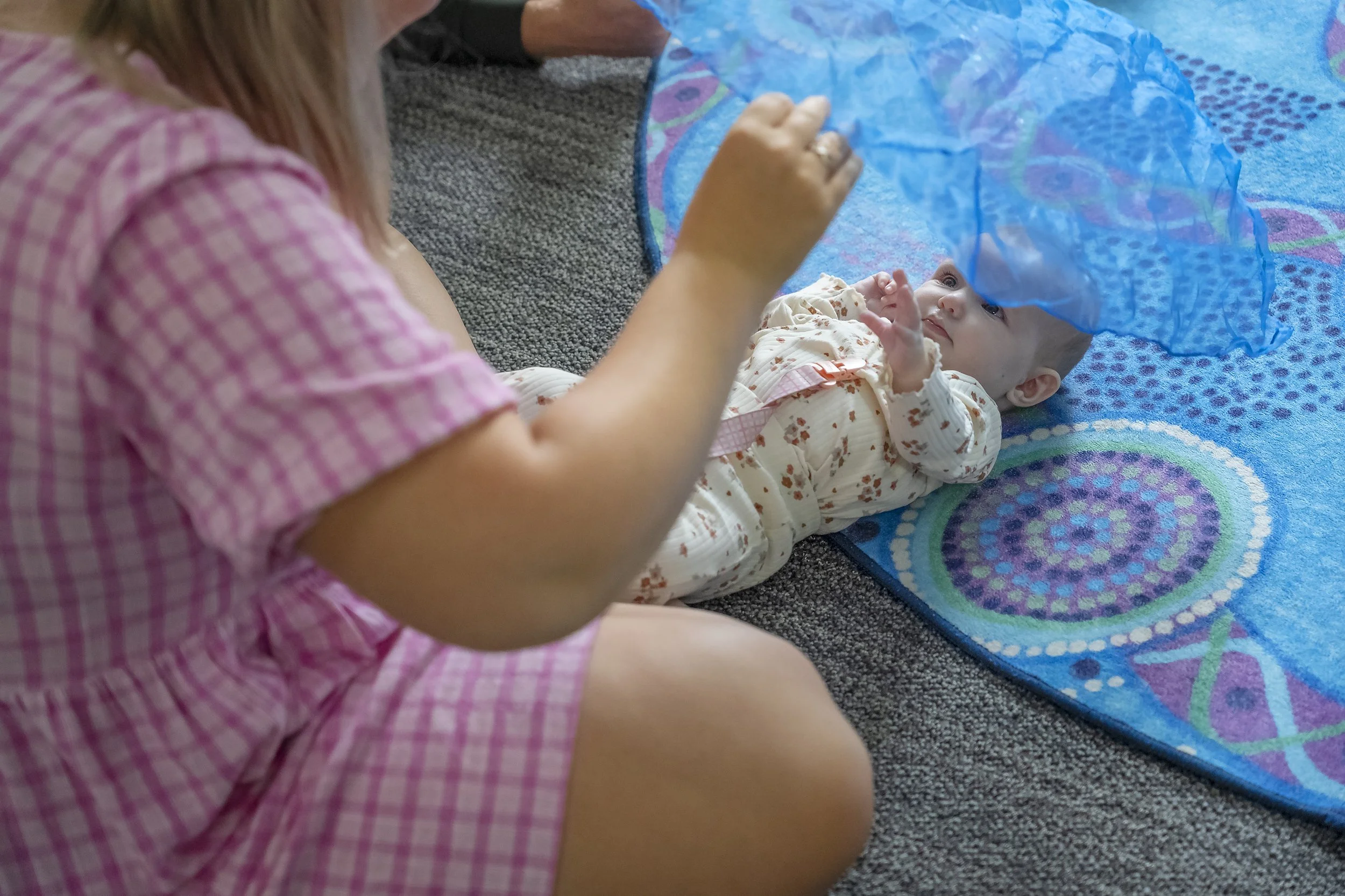 A young girl in pink checkered pajamas lying on her side, playing with a baby girl on the floor. The baby is lying on a colorful blanket and wearing cream-colored pajamas with floral print, looking up at the girl.