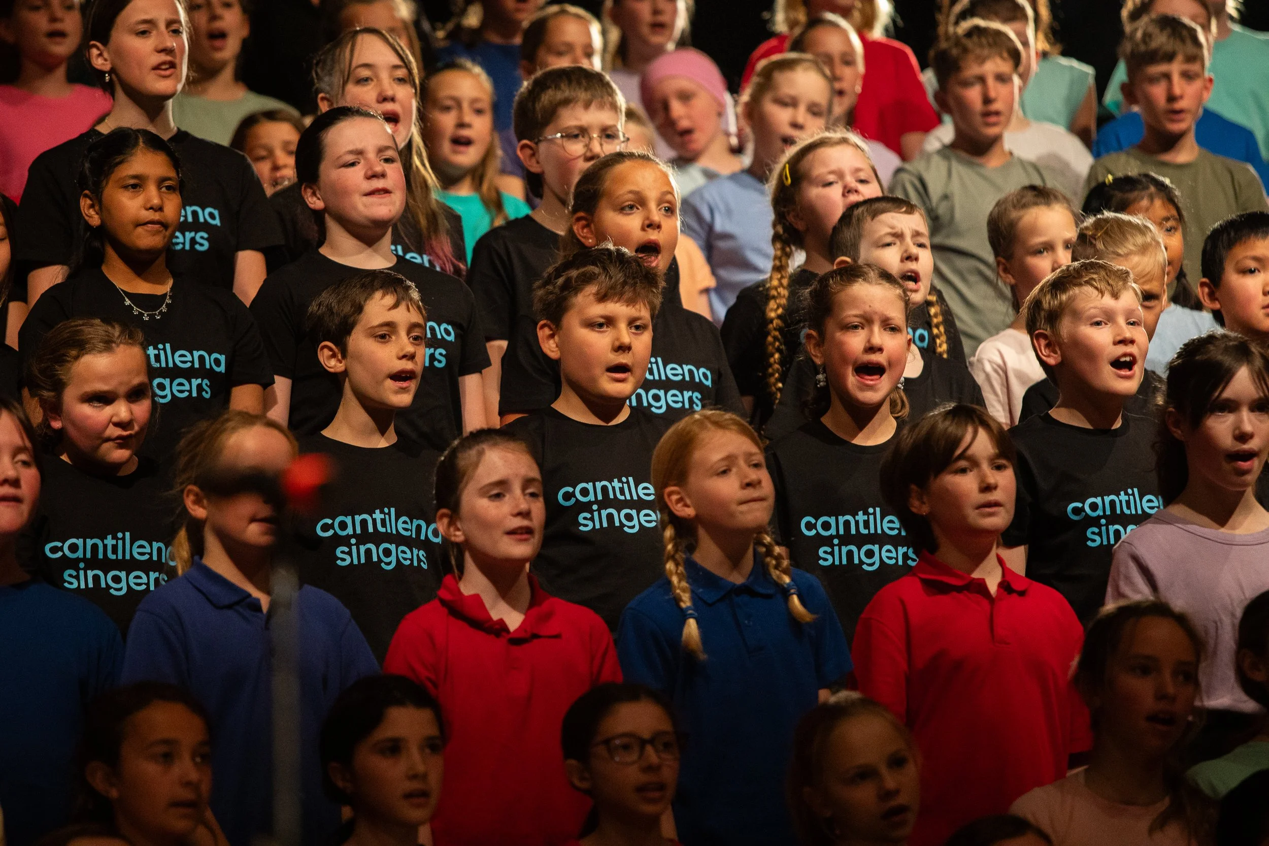Children singing in a choir on stage, wearing colorful shirts and black t-shirts with 'cantilena singers' written on them.