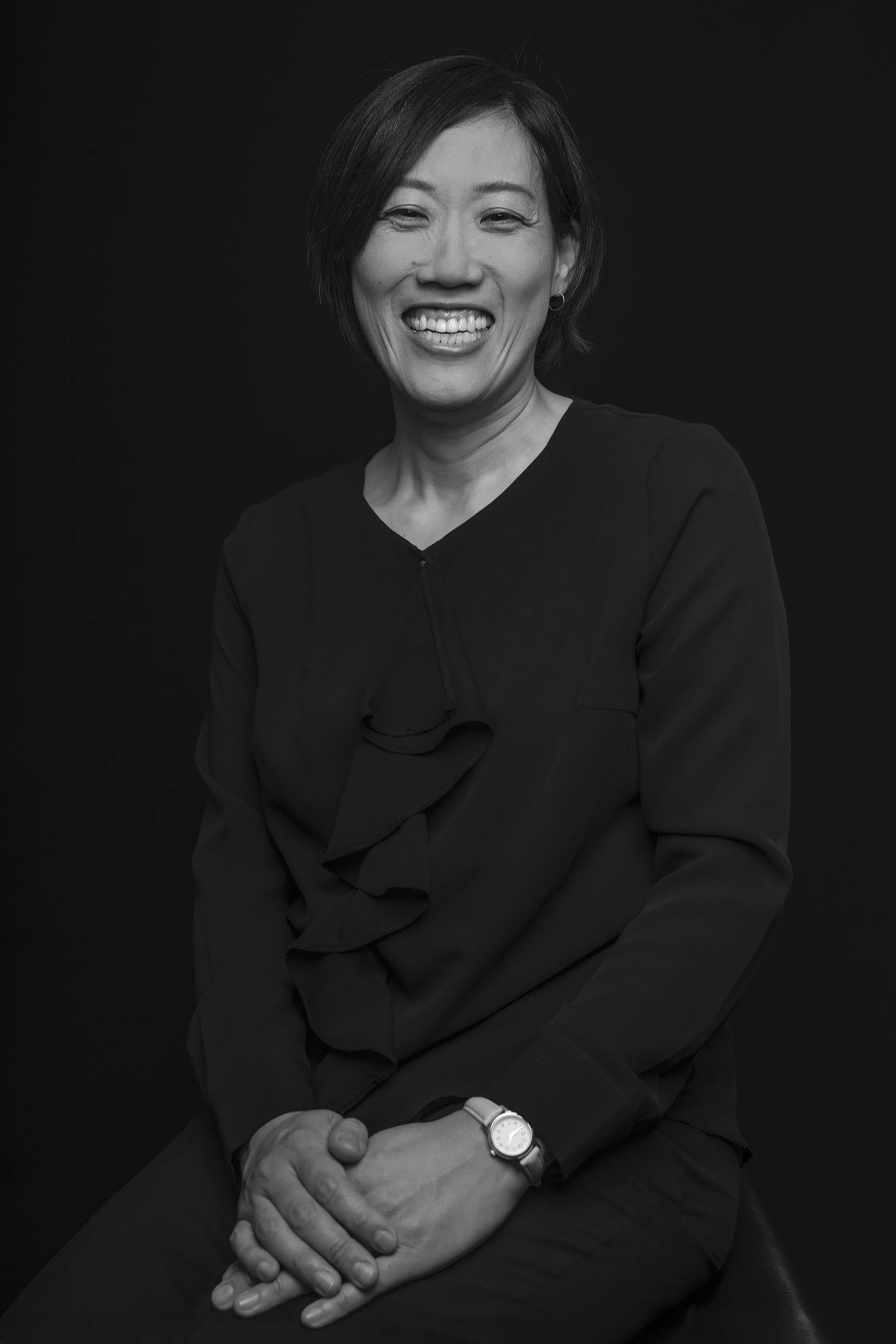 Black and white portrait of a smiling woman with short hair, wearing a dark blouse with ruffled detail, seated against a dark background.