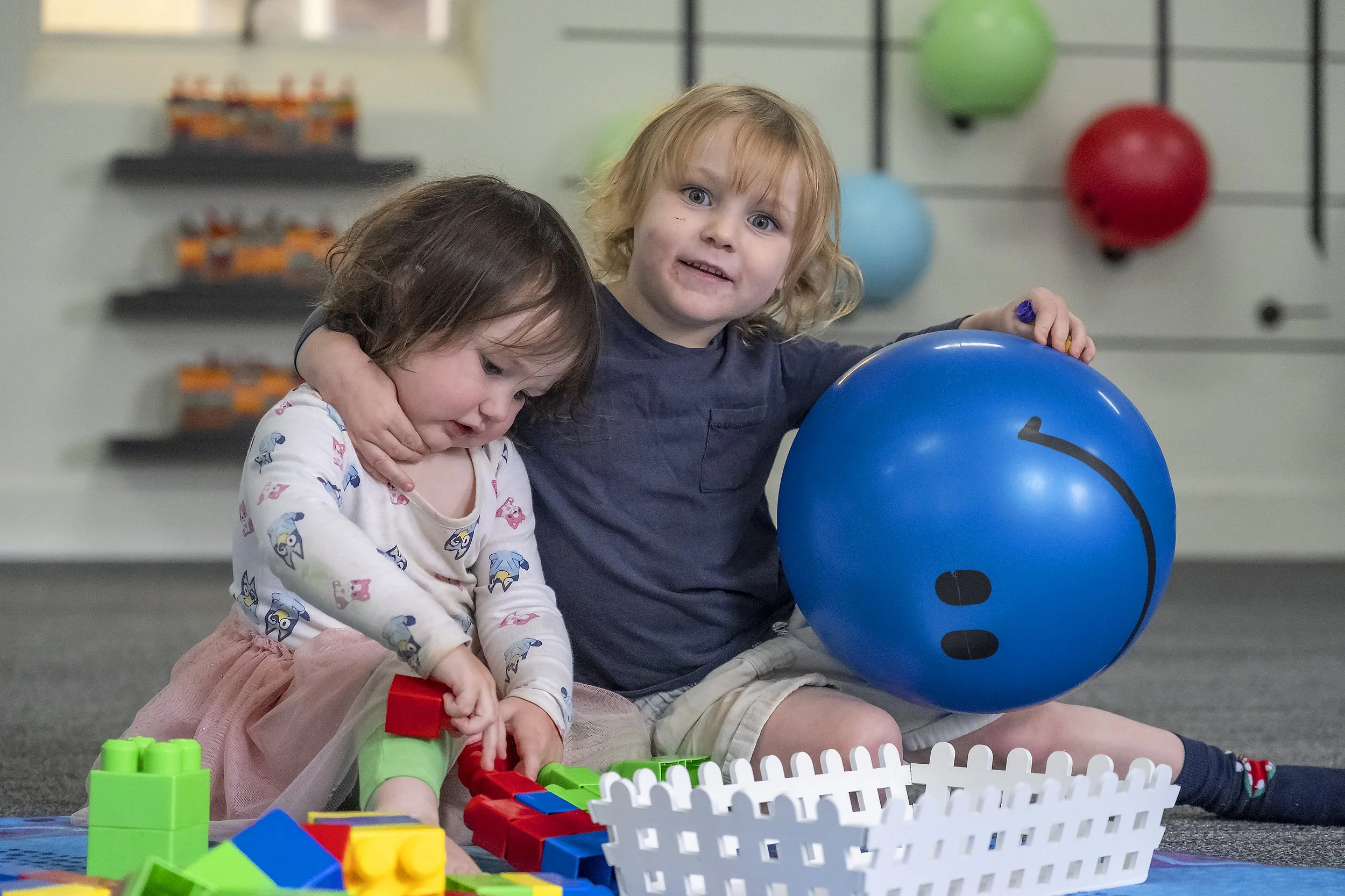 Two young children playing with building blocks and a blue exercise ball in an indoor play area.