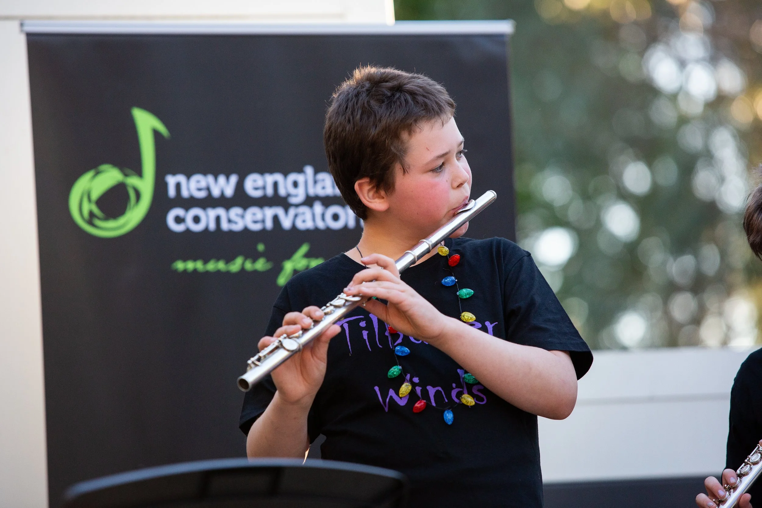A young boy playing a silver flute at an outdoor event, wearing a black shirt and a colorful string of holiday lights around his neck, with a 'New England Conservatorium' banner in the background.