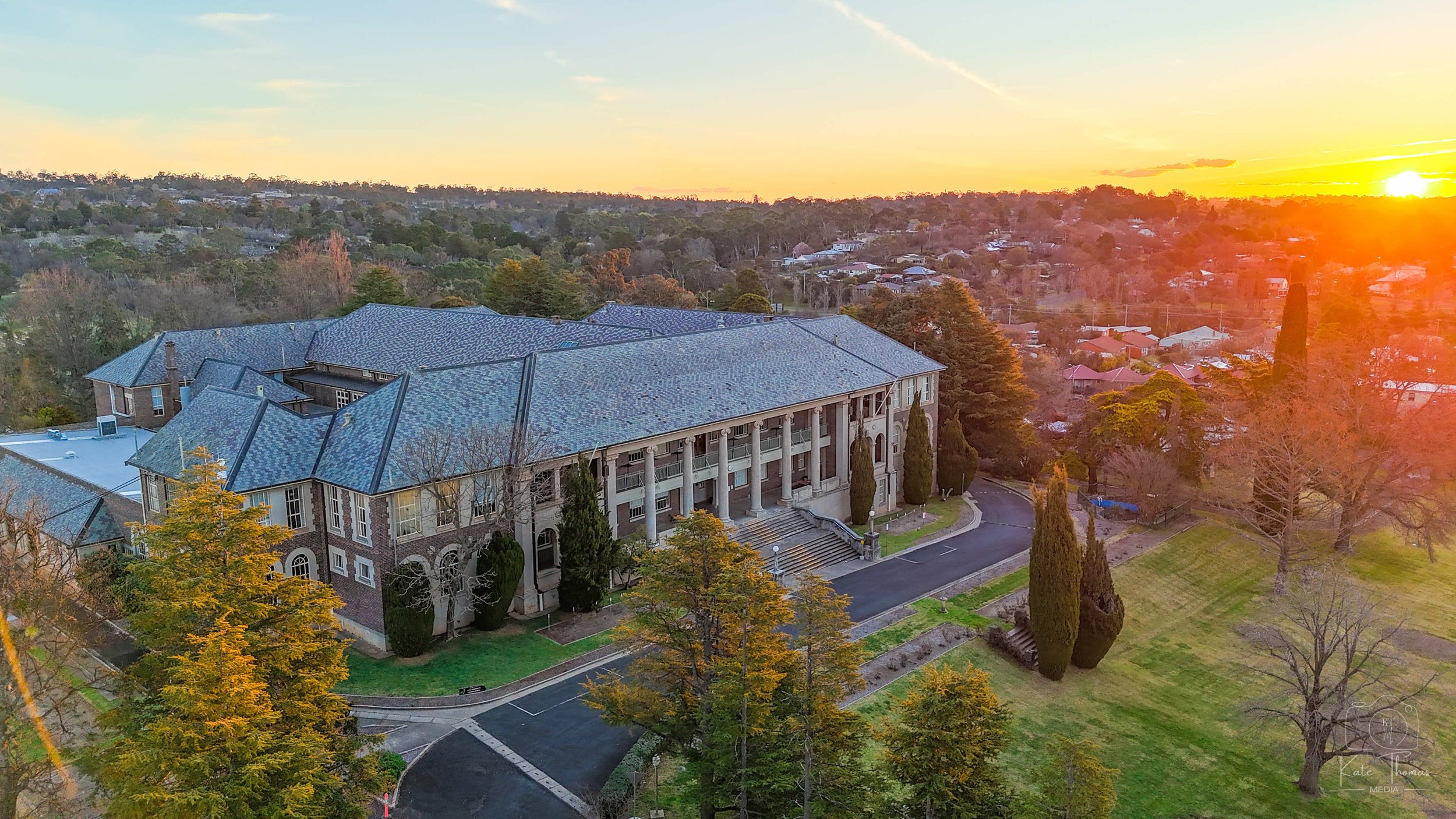 An aerial view of a large brick building with multiple stories, large columns at the entrance, surrounded by trees with fall foliage, at sunset in a suburban neighbourhood. Armidale Teachers' College, Armidale, NSW.
